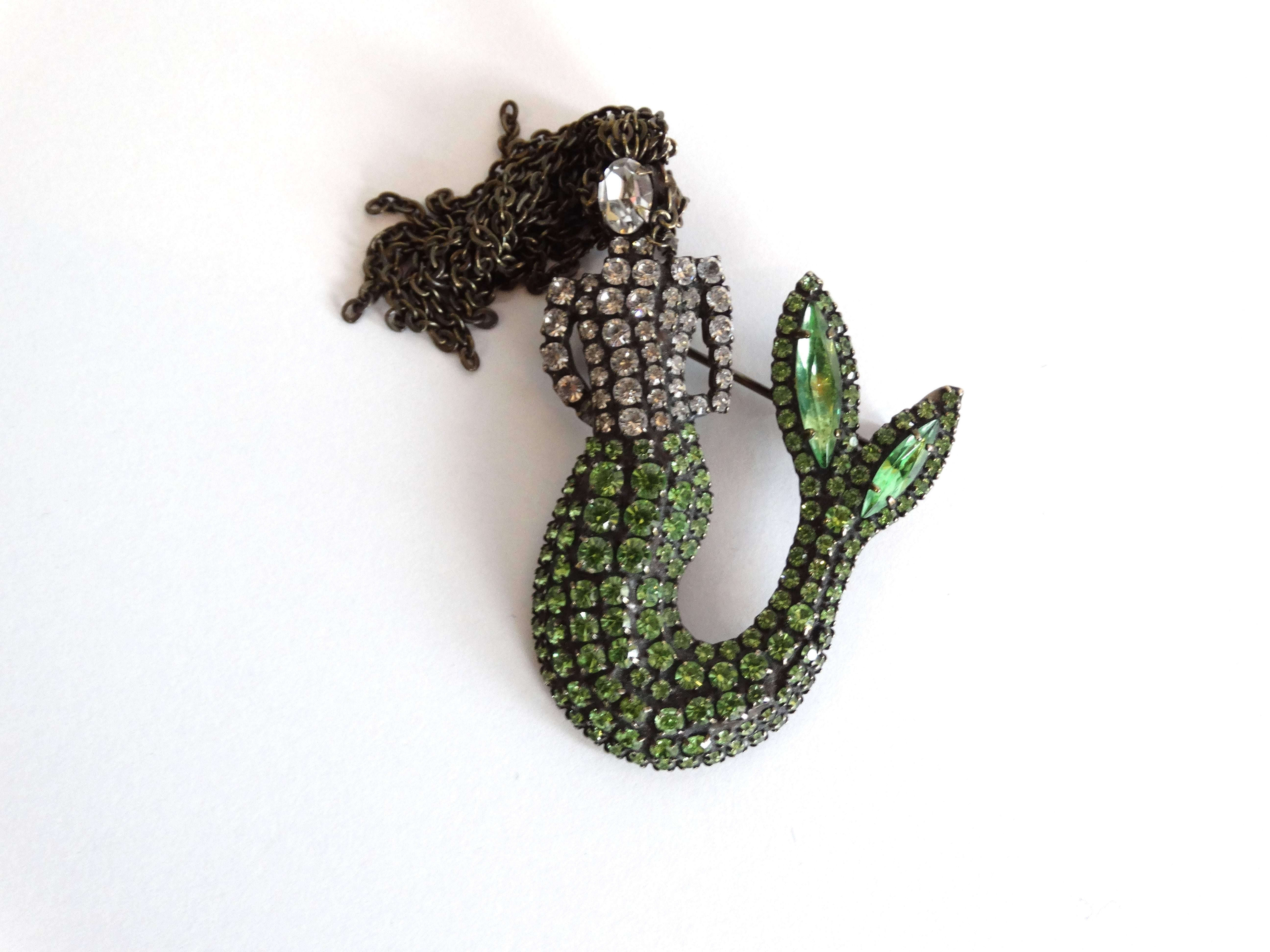 A mermaid lover's dream brooch! This alluring siren peeks out from behind her antiqued brass locks. Her upper torso is clear prong set Swarovski rhinestones and her fin is a gorgeous sea green zircon ending with 2 marquis shaped rhinestones. She