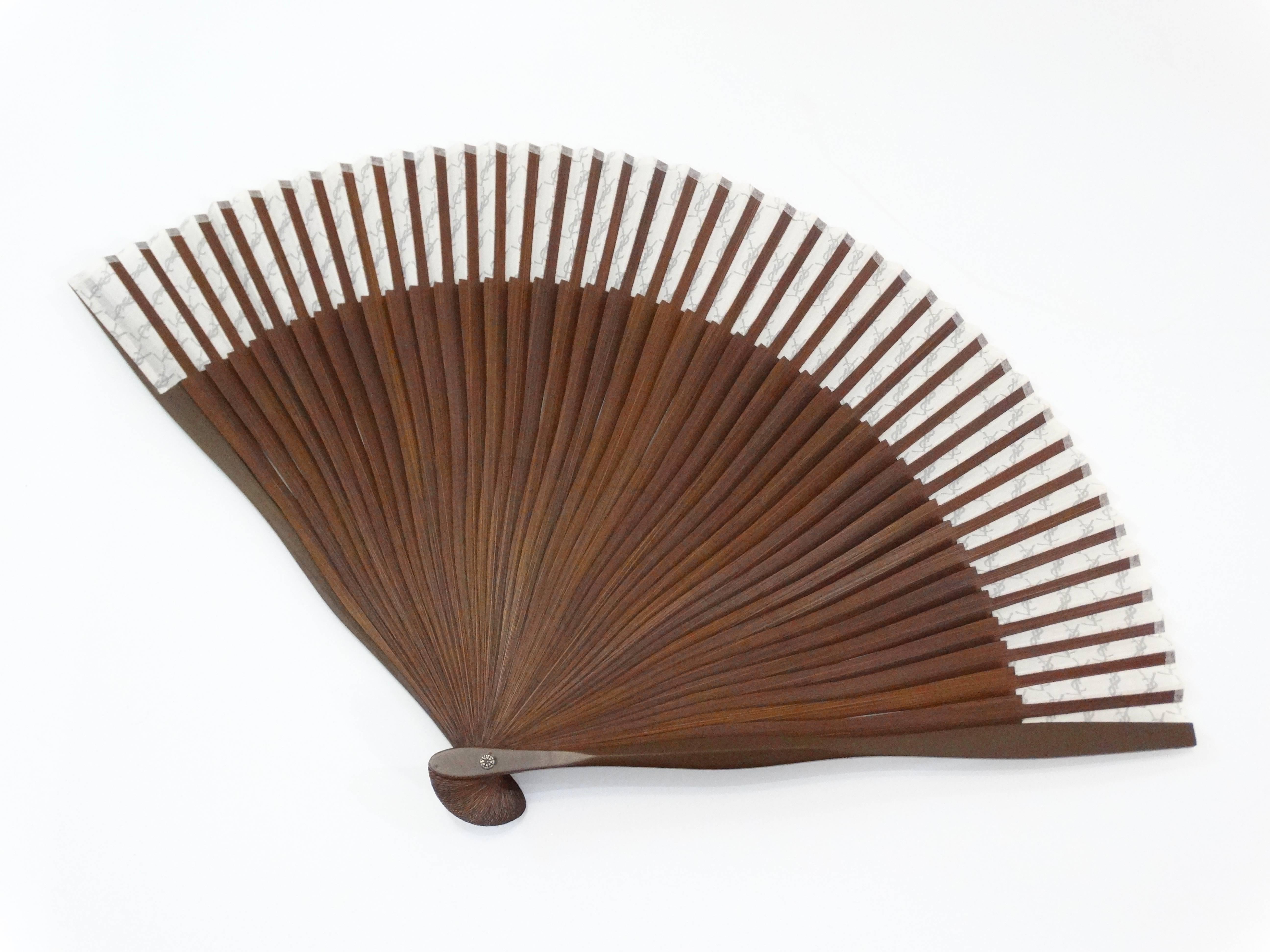 Cool off in style with our daintiest 1960s hand fan from Yves Saint Laurent! Rich brown wood handle fans out to reveal the paper YSL print in white and grey. Signed at the base of the fan with YSL signature. Comes with original ties and sheath. 