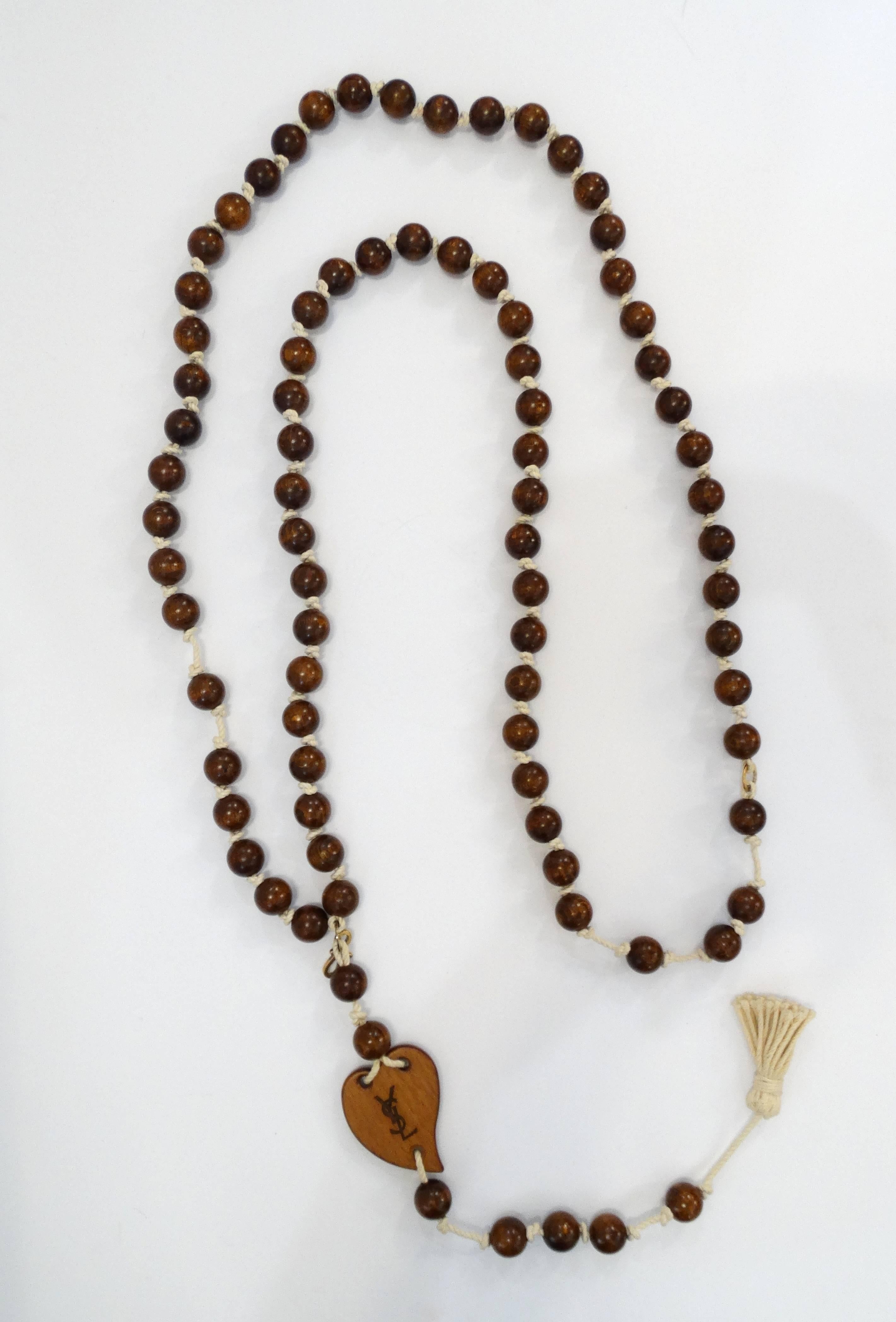 Incredible 1970s rosary style YSL necklace! Long string of brown wooden beads on thick cream colored cord. Heart shaped charm with YSL wood-burned into it. Tassel hangs from the end. Necklace can be hooked a few different ways to reduce the length.