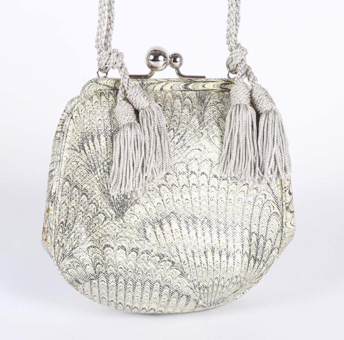 A vintage Judith Leiber minaudière. This compact purse is constructed of marbled painted lizard skin and features a decorative, silver tone kiss clasp, and a double woven shoulder strap with tassels. The interior is lined in silver satin and