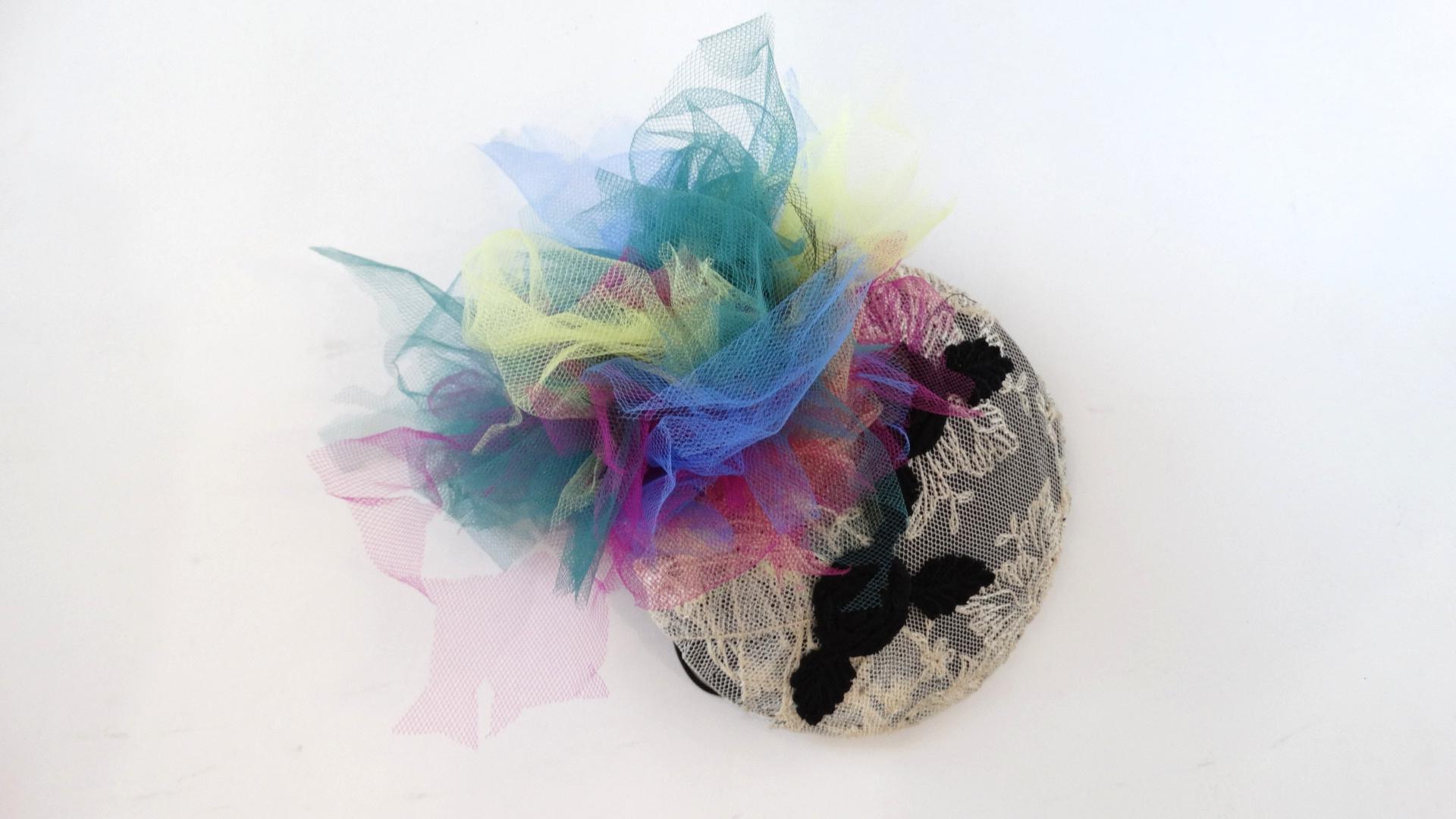 Channel your inner material girl with our amazing 1980s tulle fascinator from Selima by V! Disc shape with a cream lace overlay and black rose appliqués overtop. Spray of rainbow crinkle tulle gathered at the top. Thin black band makes it easy to