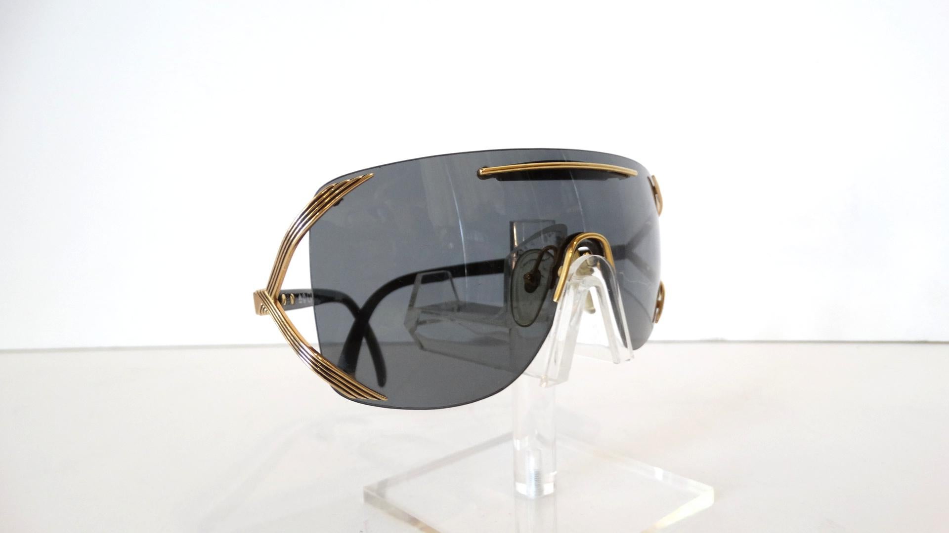A pair of 1980s vintage Christian Dior 2434 shield style sunglasses with gold tone metal accents, smoky tinted lenses, a black plastic brow bar, clear nose pads, “CD” branding at the temples and black plastic stems. They are marked to the interior