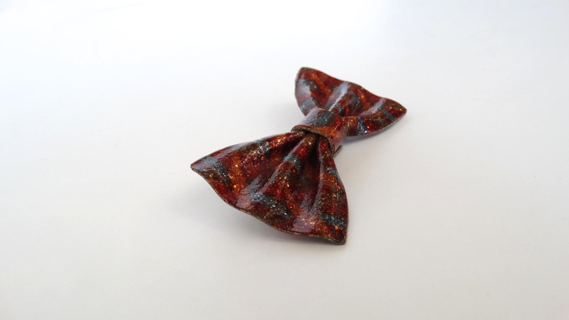 Brooches Are The New Hot Accessory This Season And We've Got You Covered! Circa 1970's, this Lea Stein bow tie brooch features a gorgeous glitter flake effect in burgundy, orange and a cool blue/grey. Shaped into the perfect structured bow tie, this