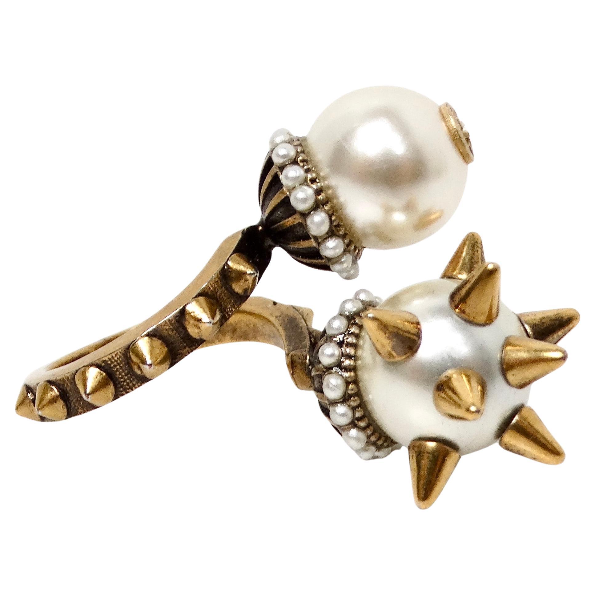 Der Gucci Faux Pearl GG & Spikes Double Ball Bypass Cocktail Ring ist ein kühnes und raffiniertes Schmuckstück, das Luxus und ausgefallenes Design nahtlos miteinander verbindet. Der offene Ring ist mit zwei auffälligen Kunstperlen verziert, die als