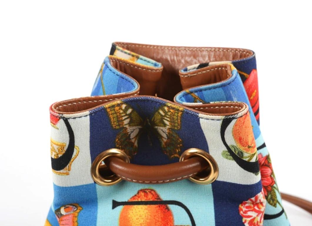 A Gucci motif print of plants, fish and fruit  multicolored canvas and leather sling backpack bag. The bag is styled with double brown leather straps interlaced through gold tone grommets to the cinch closure and brown leather to the base. The