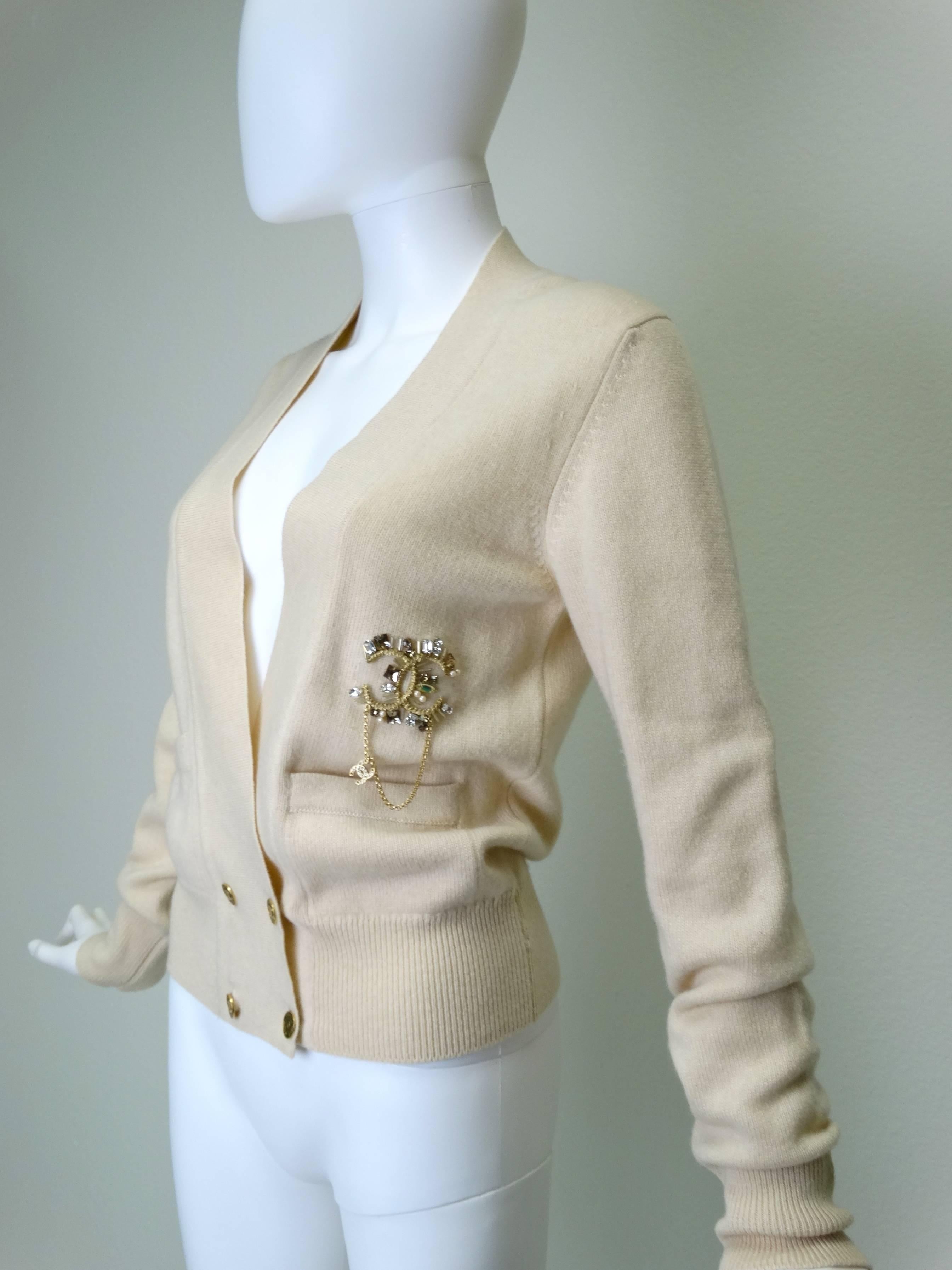 A Chanel 2006A 100% cashmere deep v-neck sweater with double breasted buttons. Sweater is crème with a beautiful encrusted crystal CC logo on the left side. CC emblem has faux crystals, pearls and rhinestones with a gold chain hanging down which
