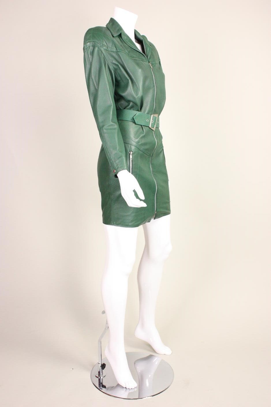 Super sexy Thierry Mugler green leather mini dress dates to the late 1980's through the early 1990's.  It features a zippered front closure, zippered pockets at hip, and is fully lined. Padded shoulders. Detached belt with silver-toned