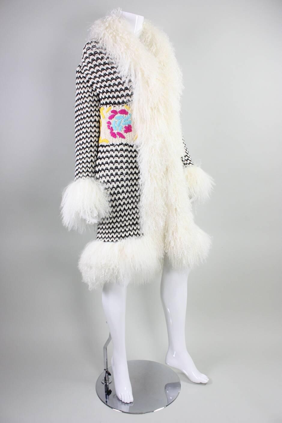 Cardigan/coat from Escada Sport likely dates to the 1990’s to 2000’s.  It is made of black and cream wool yarn that is knit in a zigzagged stripe pattern.  Wide waist band has a brightly colored floral motif.  Cream Mongolian lamb wool trim.  Center