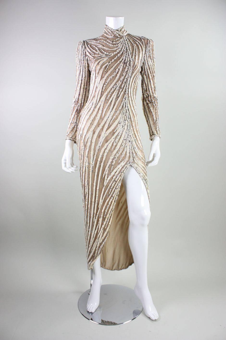 Vintage evening gown from Bob Mackie dates to the 1980's and is made of nude stretch netting that is adorned with sinuous bands of silver bugle beads, cream sequins, and pearls.  Gown has long sleeves with zippered cuffs, a mock neck, and is fitted