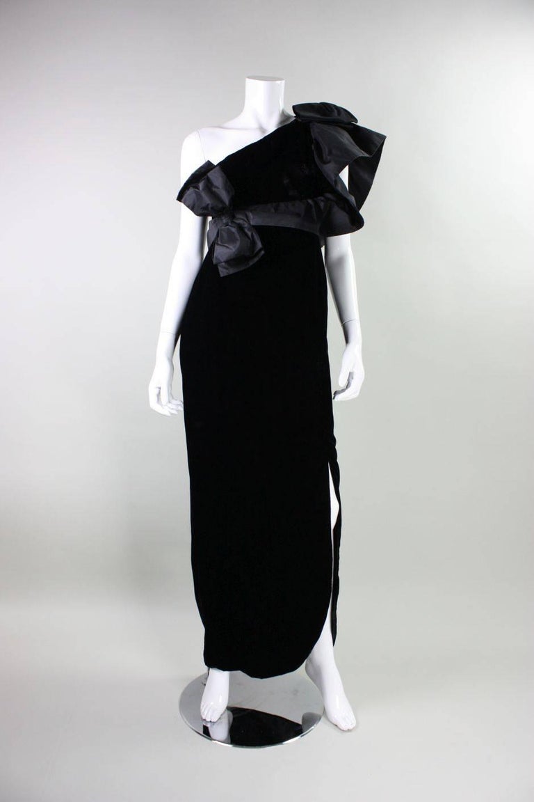 Vintage gown from Adolfo dates to the 1980's and is made of jet black velvet with black silk taffeta accents. Diagonal asymmetrical neckline with oversized ruffled shoulder. Slit in hem at left calf. Center back diagonal zipper.  Fully