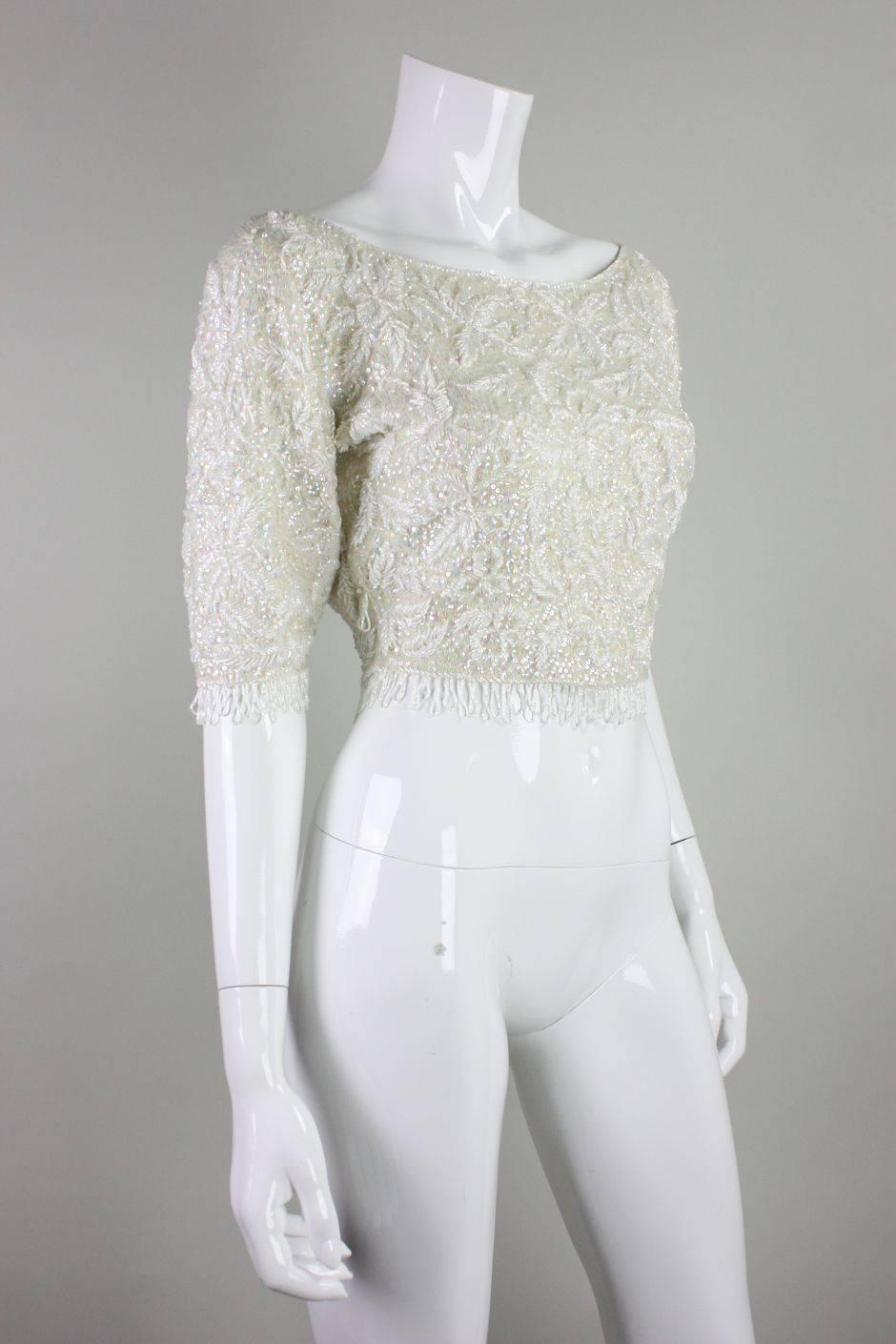 Vintage cropped sweater from Jo-Ro Imports retailed at I. Magnin and is made of cream-colored wool.  Sequins are iridescent and are accented with white bugle beads that are worked in a floral pattern.  Beaded looped fringe trims the cuffs and