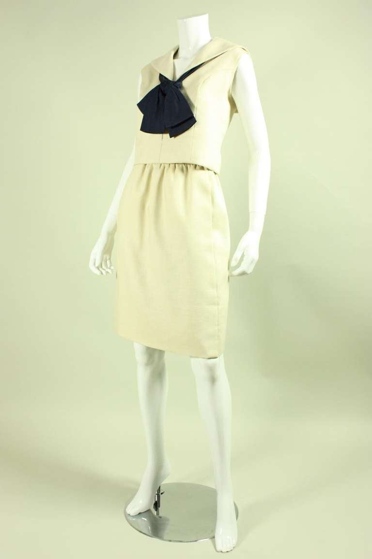 Two piece vest and skirt ensemble from Norman Norell dates to the 1960's. It is made of cream linen with a navy silk faille bow that is detachable. Vest has a v-neck, middy collar, and center front zipper. Straight skirt is gathered all around