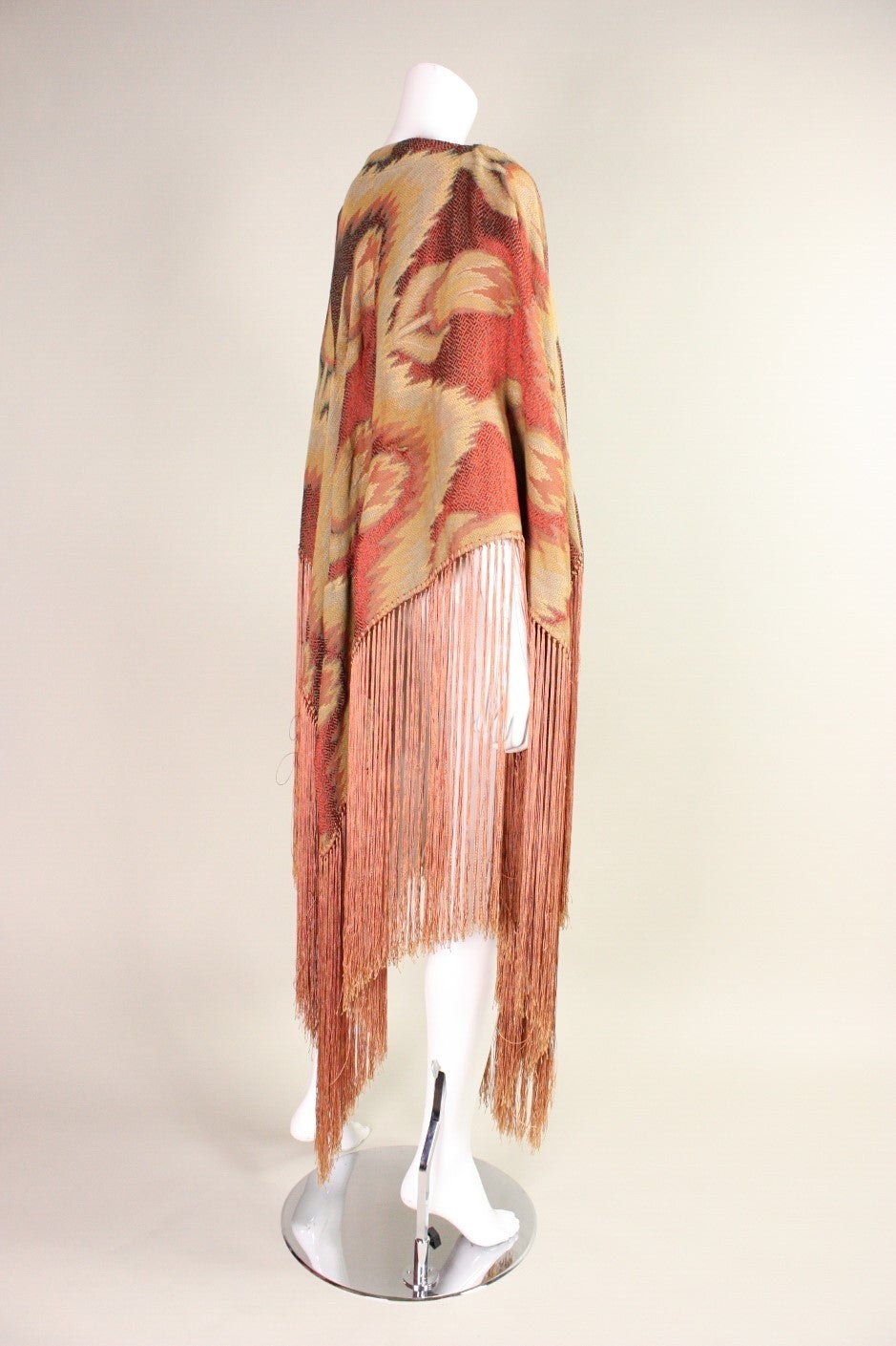 Vintage cape likely dates to the 1970's through 1980's and is made of a dusty rose jacquard fabric with a floral ikat-like print.  Rows of long fringe hang down from the hem.  Unlined.  No closures. Piece is signed along the leather-lined neckline;