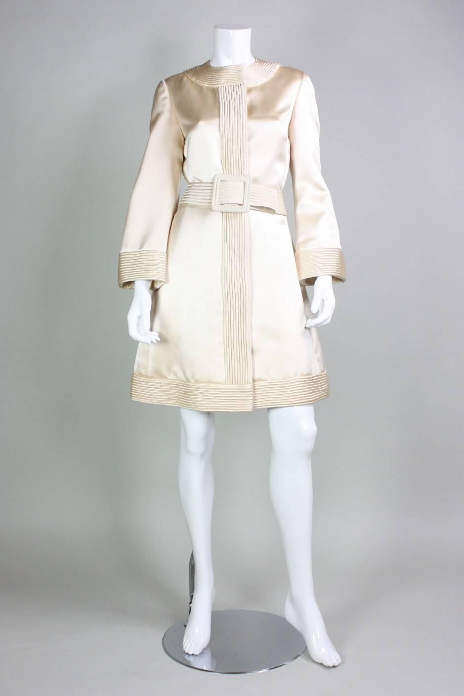 Space-age dress from Donald Brooks dates to the 1960's and is made of cream-colored silk satin.  Wide bands of fabric are concentrically stitched around neckline, at cuffs, down center front, and around the hem.  A-line skirt.  Wide bell sleeves. 