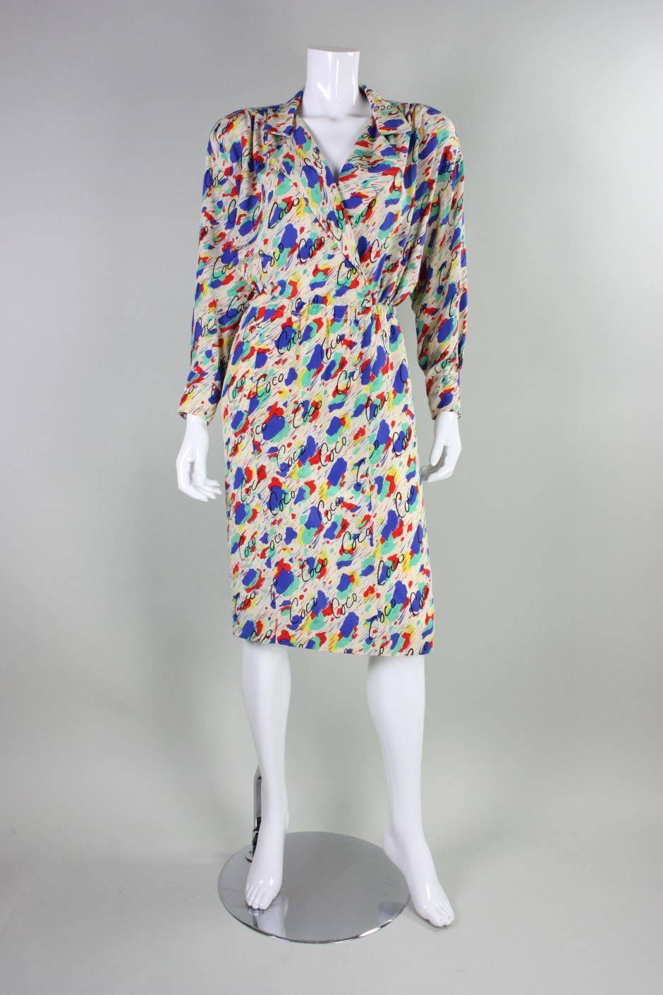 Vintage dress from Chanel is made of off-white silk that features a polychromatic painterly print with cursive Coco written throughout. Crisscrossed bodice has a v-neck, notched lapel, and long sleeves with buttoned cuffs.  Straight skirt has a 