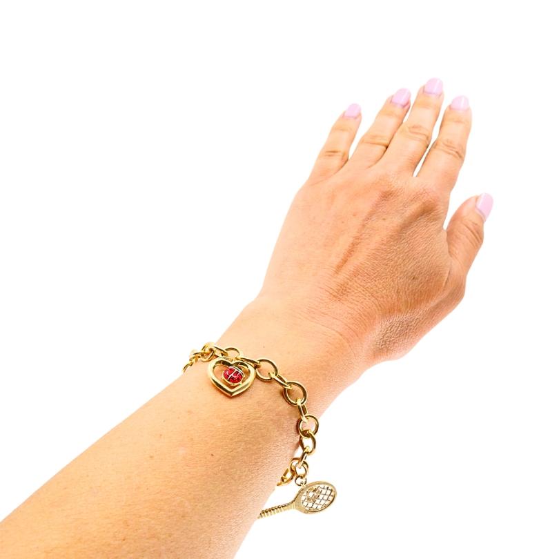 Una pulsera de eslabones dorada con dos divertidos colgantes en una clásica cadena de eslabones ovalados. La pulsera está hecha de eslabones uniformes con un acabado liso y un cierre de langosta seguro. Un amuleto es una raqueta de tenis de oro con