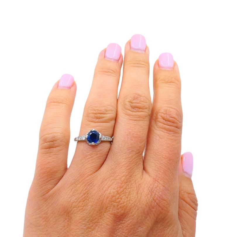 Dieser Art-Déco-Ring ist aus Platin gefertigt und trägt in der Mitte einen runden, tiefblauen Saphir, der von vier Zacken gehalten wird. Der Saphir wird umrahmt von einem zarten Heiligenschein aus  Diamanten, die sich an den Schultern des Bandes
