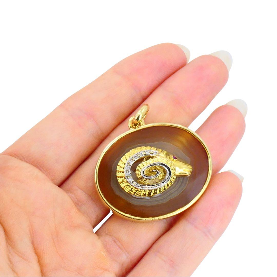 Dieser Anhänger aus 18-karätigem Gelbgold zeigt einen skulpturalen Widderkopf, der auf einer natürlichen Achatplatte ruht. Der ovale Achat-Sockel weist eine satte braune und karamellfarbene Bänderung auf, die einen starken Kontrast zur polierten
