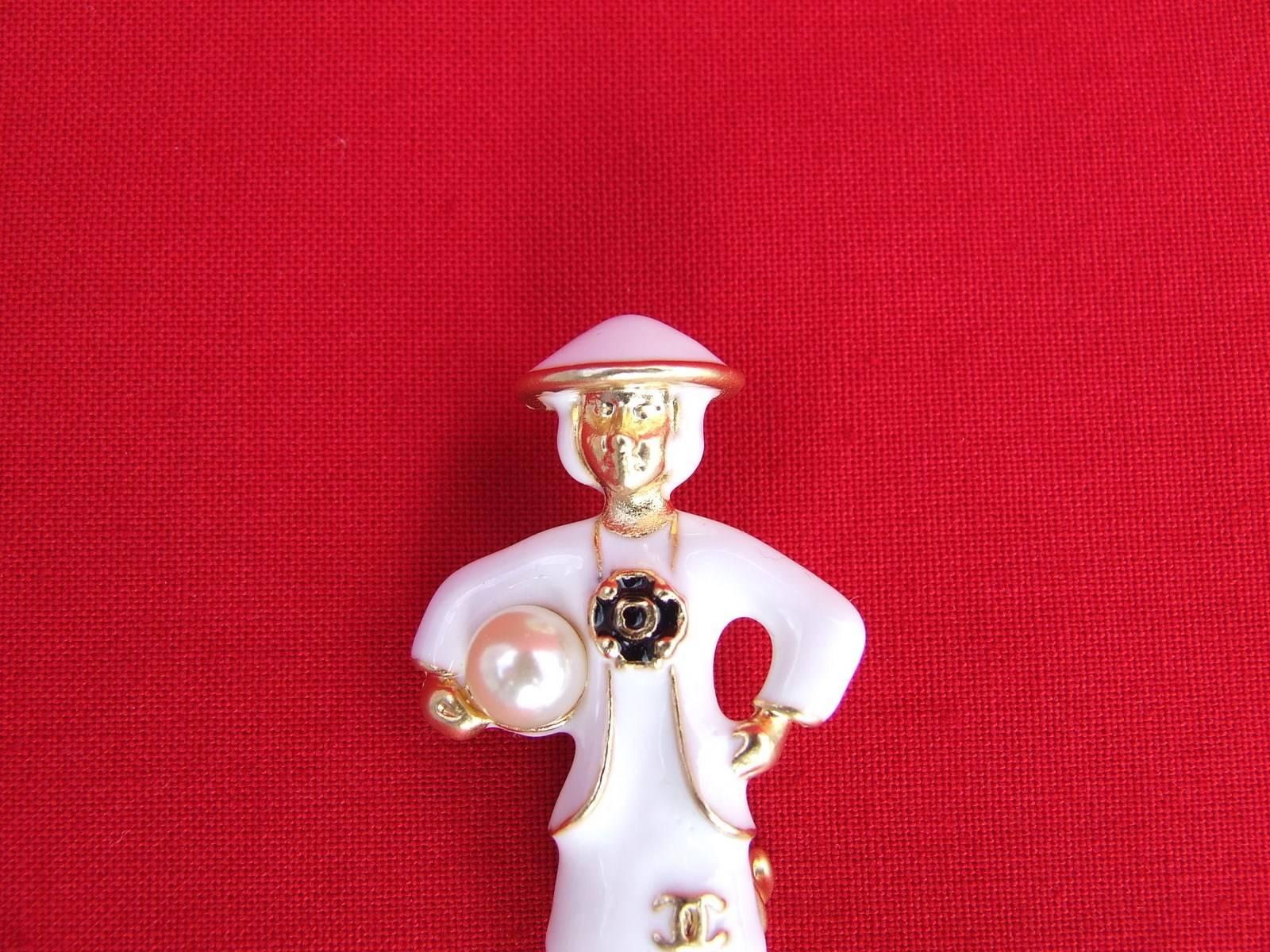 Beautiful Authentic CHANEL Brooch

Made of golden Metal, dress and hat in white Enamel

Represents Mademoiselle Coco Chanel in a white dress, holding a false ivory pearl under her right arm

Black Camelia in the middle, and cc logo in golden metal