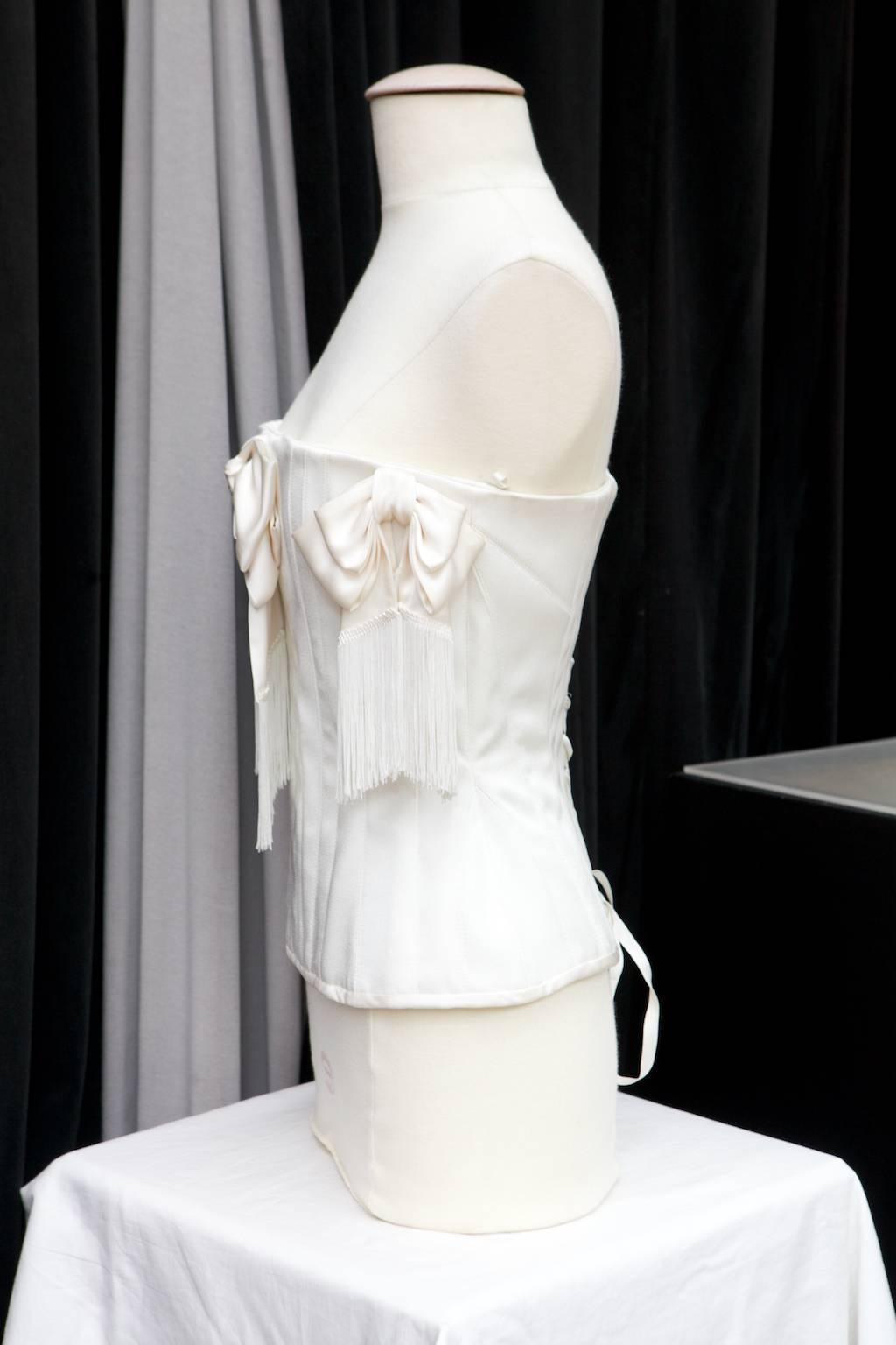 JOHN GALLIANO (Made in France) Corset in white satin in a victorian style decorated with three large bows with dropping fringes around the chest.

The bustier fits perfectly the waist thanks to semi flexible reinforcements and a lace work at the
