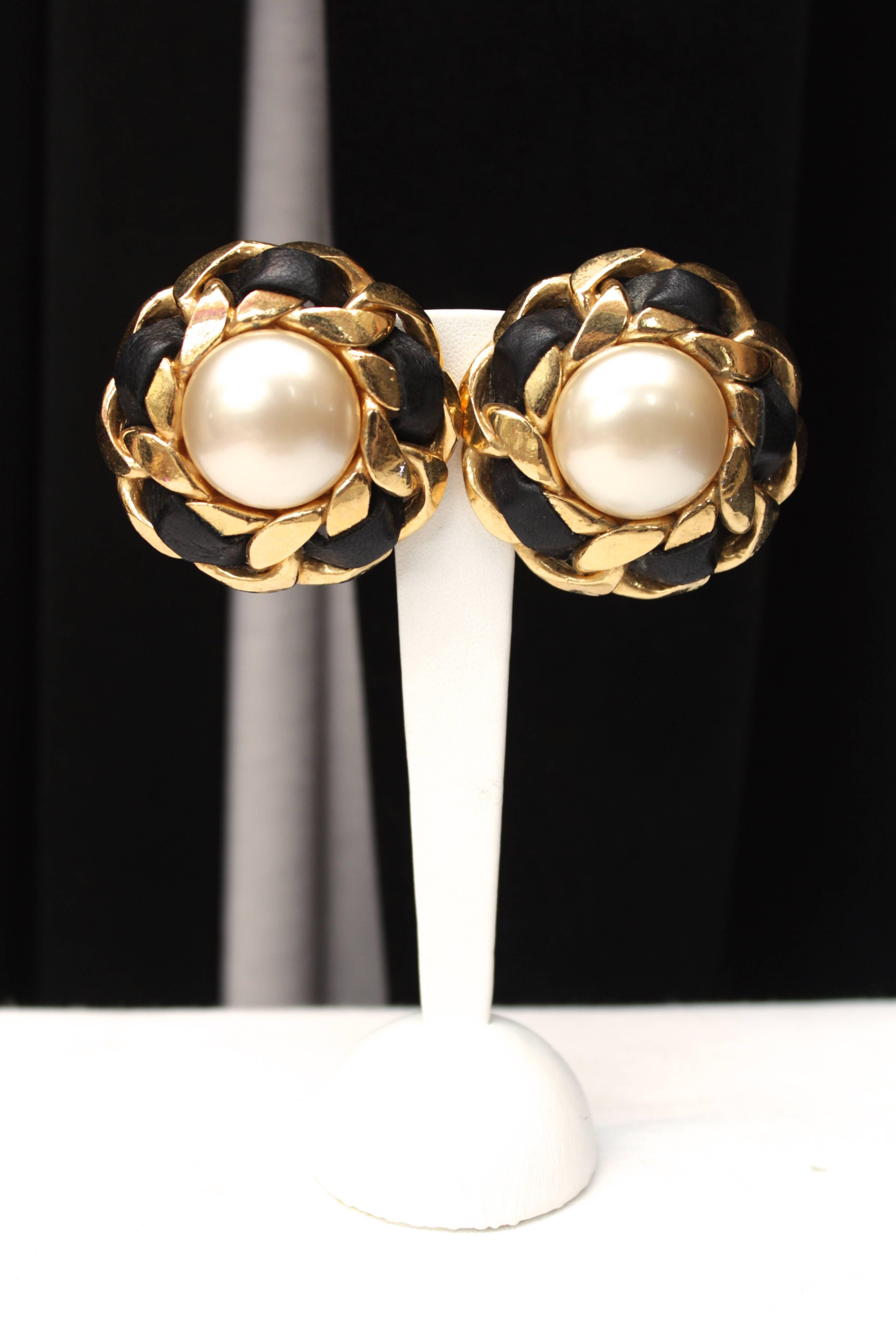 CHANEL (Made in France) Large clip on earrings composed of a curb gilt rigid chain interwoven with a black leather strap and centered with a faux pearl cabochon.

The earrings are signed on the back. 

They date from the late1980s / early