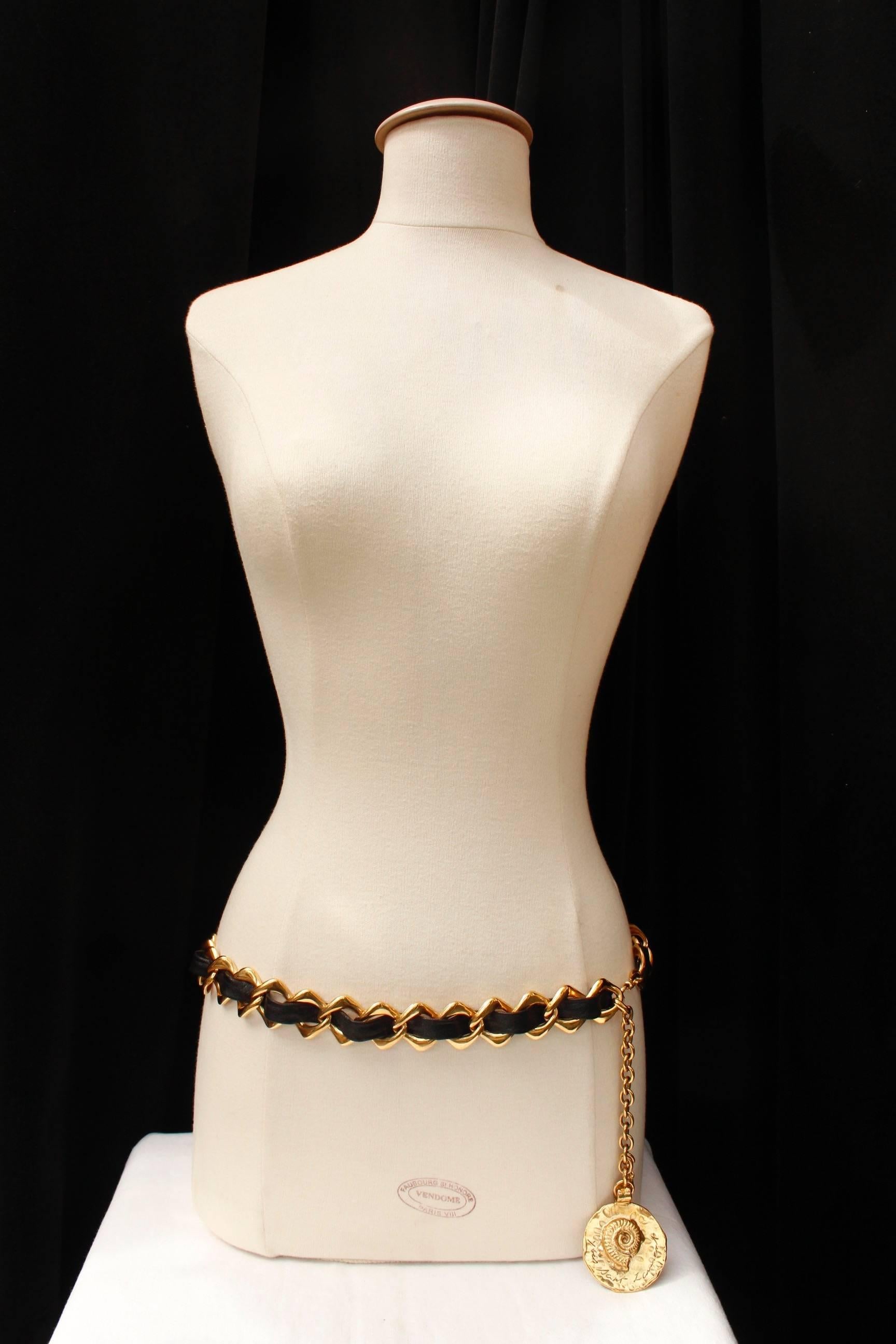 YVES SAINT LAURENT (Made in France) – Belt composed of a wide openwork gilded metal rhomboid chain entwined with a wide black leather strip. It is adjustable by means of a long gilded metal chain ending with a round hammered pendant decorated with a