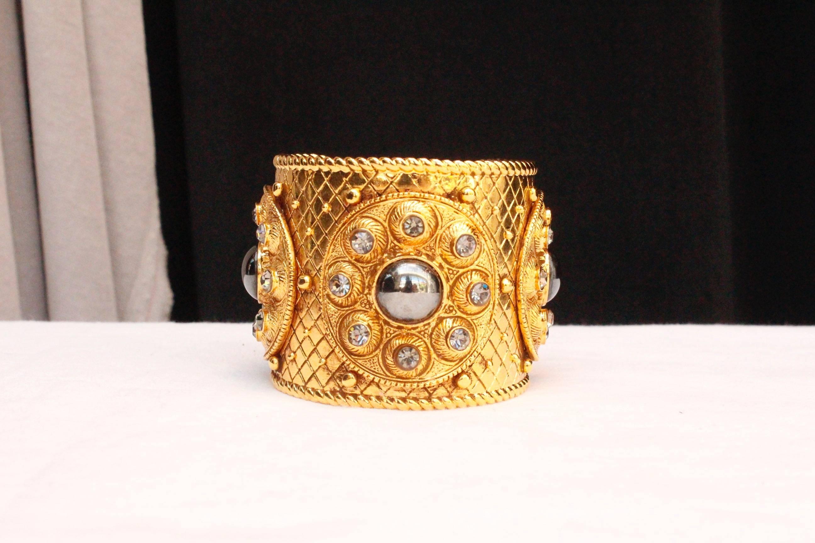 CHRISTIAN DIOR BOUTIQUE – Cuff bracelet composed of chiseled gilded metal decorated with three circles paved with rhinestones and a centered grey cabochon.  Signed inside.

Circa 1990.

Wrist circumference 17.5 cm (7 in); Opening 3 cm (1.25 in);