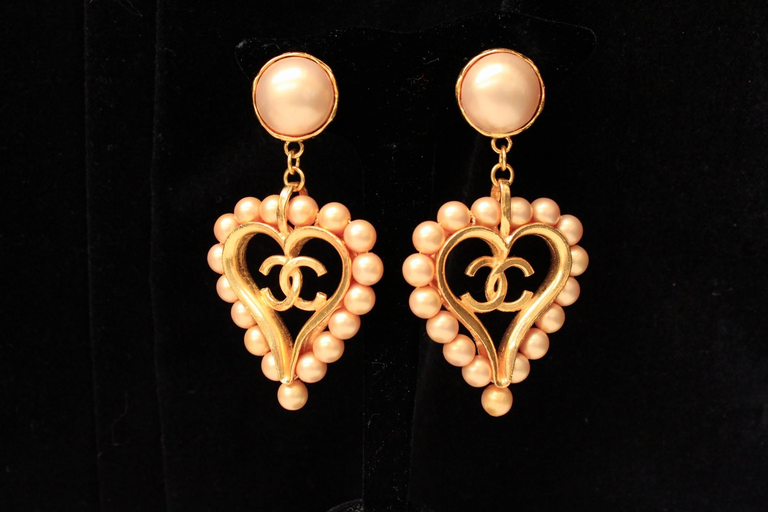 CHANEL (Made in France) Drop earrings composed of a gilded metal clip set with a gold tone pearly glass paste cabochon. The drop part represents an openwork gilded metal heart with CC logo, surrounded with small golden beads.

Signed at back.

2cc8