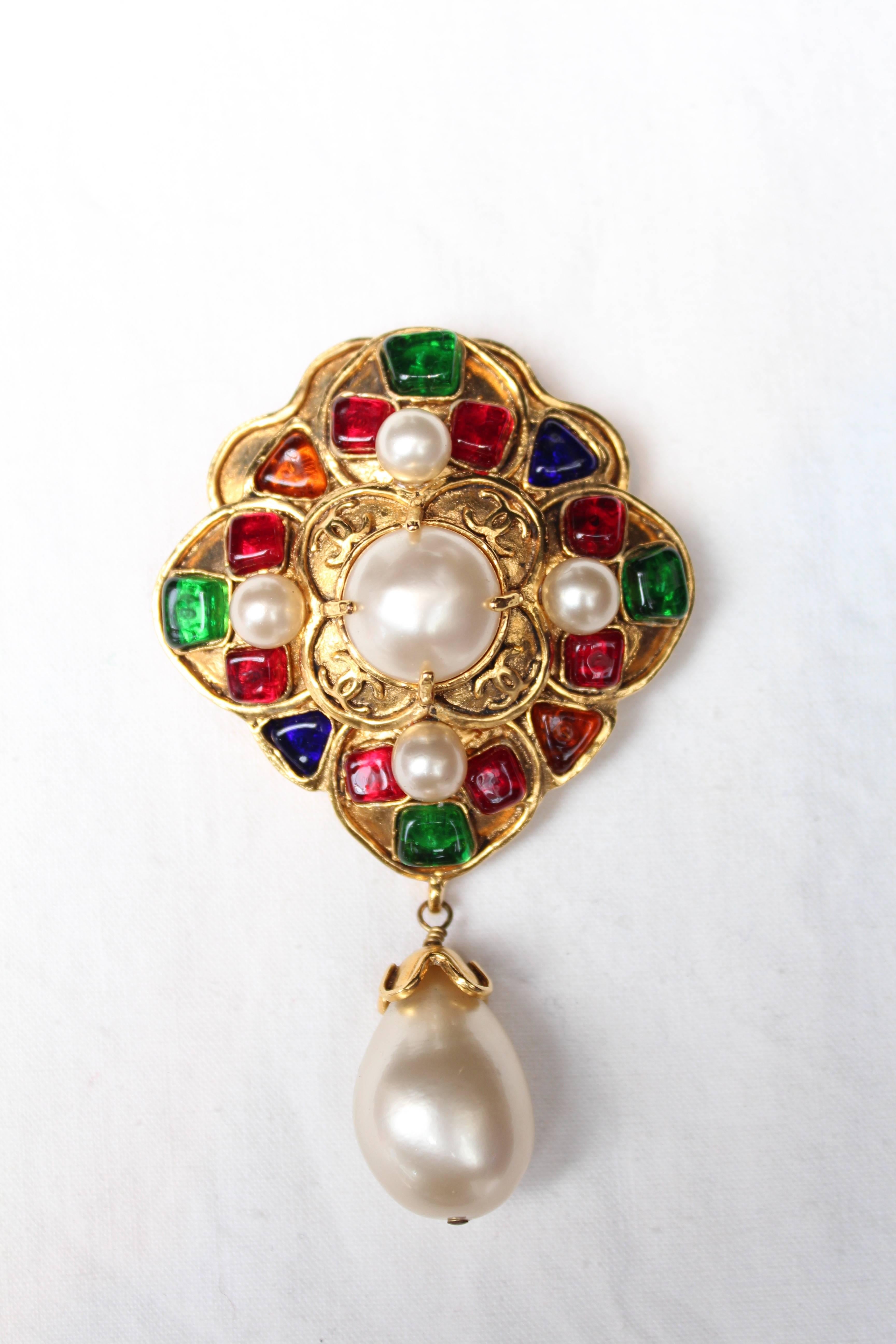 CHANEL (Made in France) Beautiful hammered gilded metal brooch representing a stylized cross paved with ruby, sapphire and emerald glass paste cabochons. A pearly cabochons decorates the center of the brooch. A pearly teardrop topped with a gilded
