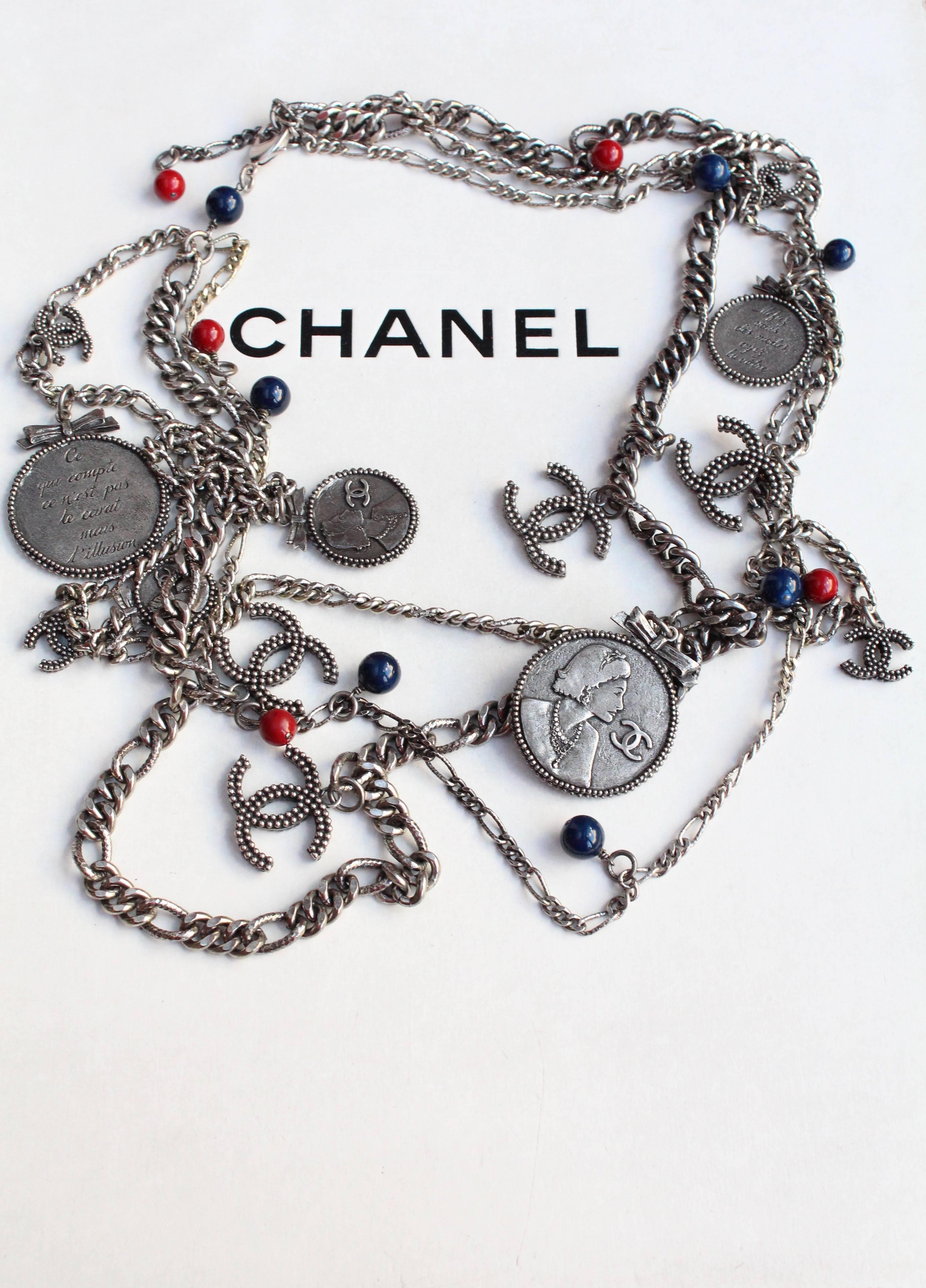 CHANEL (Made in France) Long necklace comprised of three strands of rhodium plated metal chain with dangling silver tone medals topped with a small bow, silver tone CC logos and red and blue glass beads.

The round medals feature on one side a