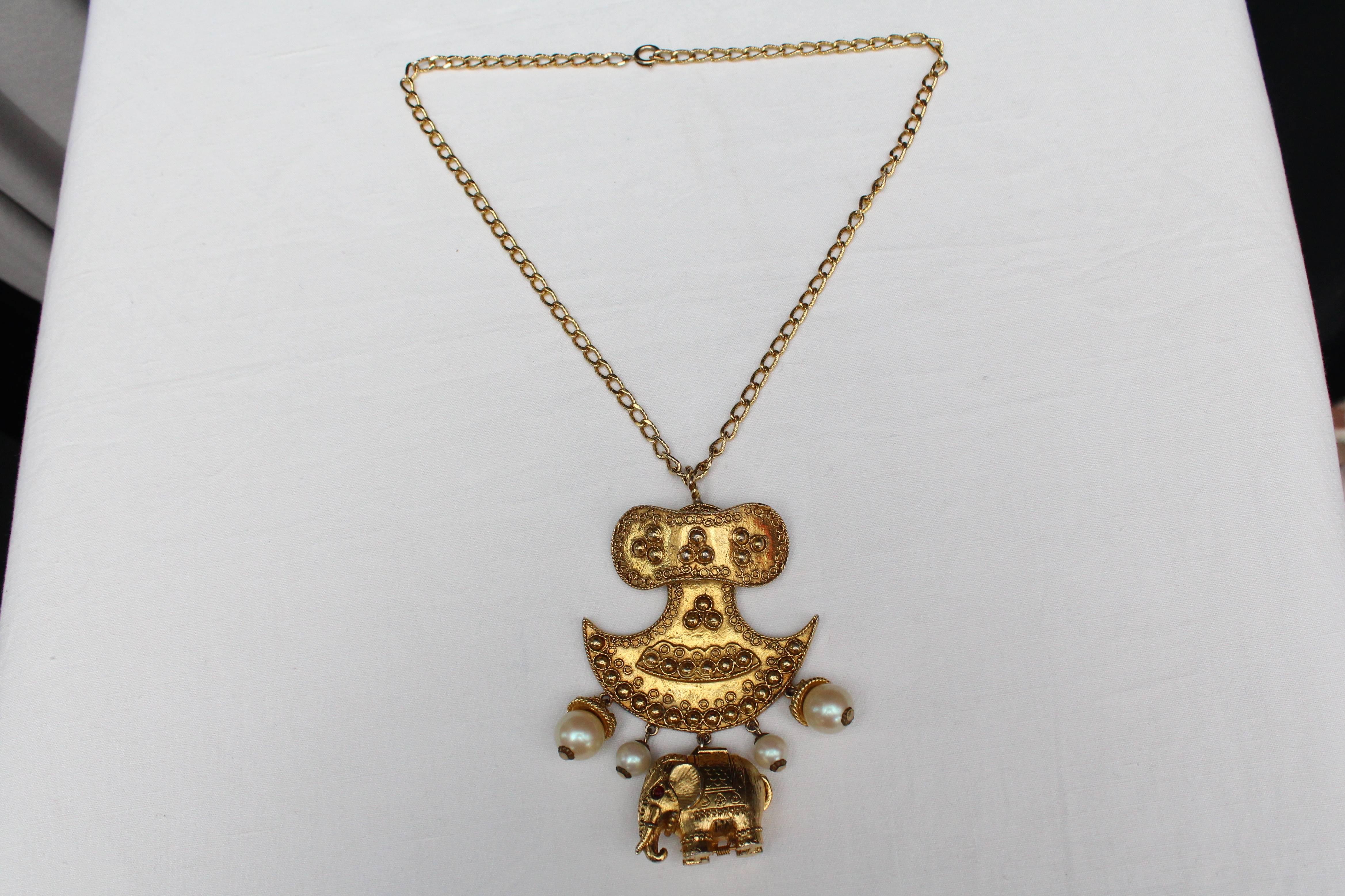 KENNETH LANE gilt metal necklace in a etruscan style composed of a chain holding a large golden metal pendant engraved holding a series of faux-pearls and a gold metal elephant paved with two red rhinestones in the place of the  eyes. 
The elephant
