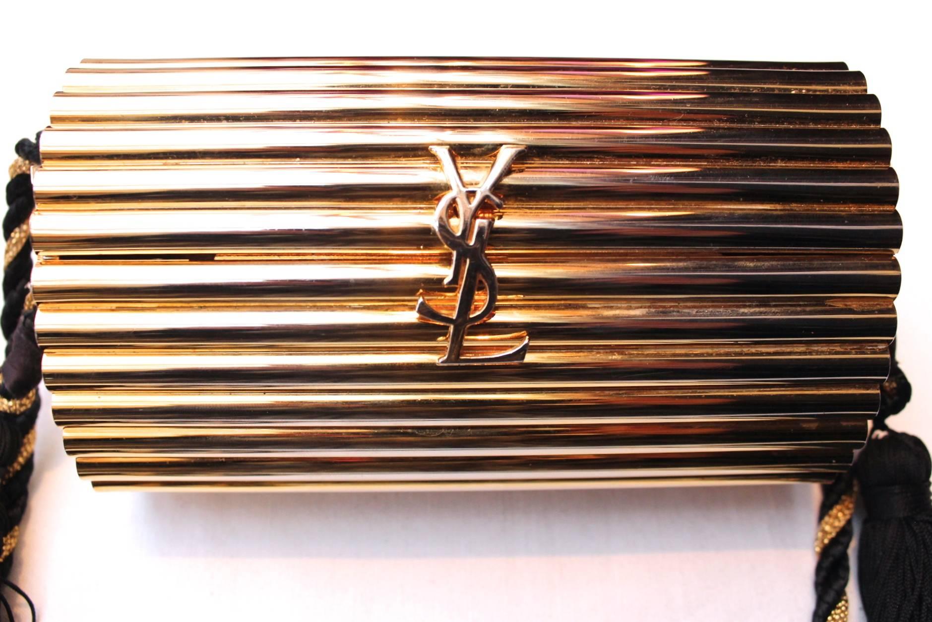 YVES SAINT LAURENT Gilt metal evening clutch featuring a chiseled and ribbed textured oval box, a large logo front detail, a black and gold braided shoulder strap and two black passementerie tassels. The purse is lined in gold leather

A must have