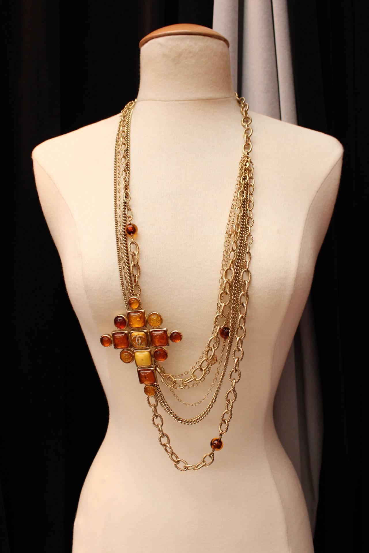 CHANEL (Made in France) Large necklace composed of several strands of chains in light gilt metal adorned with a cross in gilt metal paved with glass paste square and round cabochons in orange, amber and yellow colors. 

The center cabochon