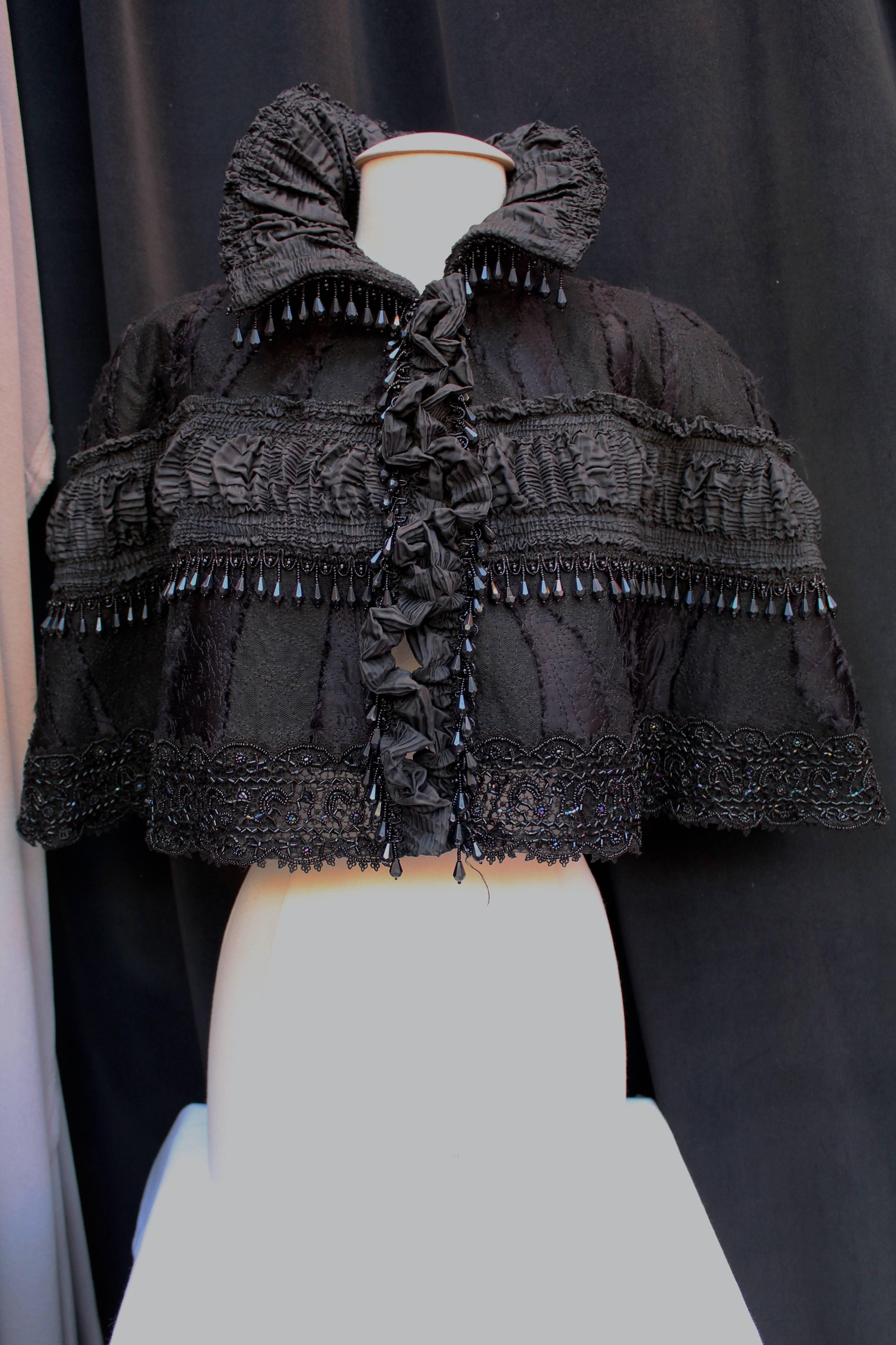 CHRISTIAN LACROIX (Made in France) Short cape in a victorian style composed of black synthetic fabric embroidered with black lace and black beads. 

The cape is also adorned with torn straps of black fabric and a horizontal strip of pleated black