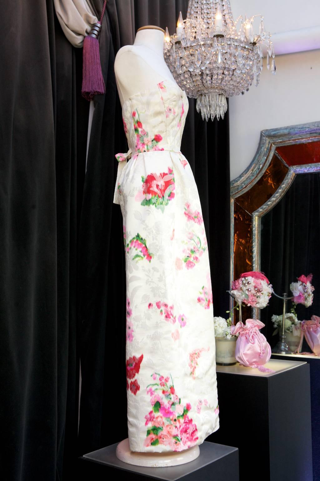 NINA RICCI HAUTE COUTURE by Jules-François Crahay. Long evening dress made of off-white satin brocade with fuschia flower print. The straight skirt features front and rear darts. A tonal ribbon is fastened to the waist. Back zip closure. 
White tag