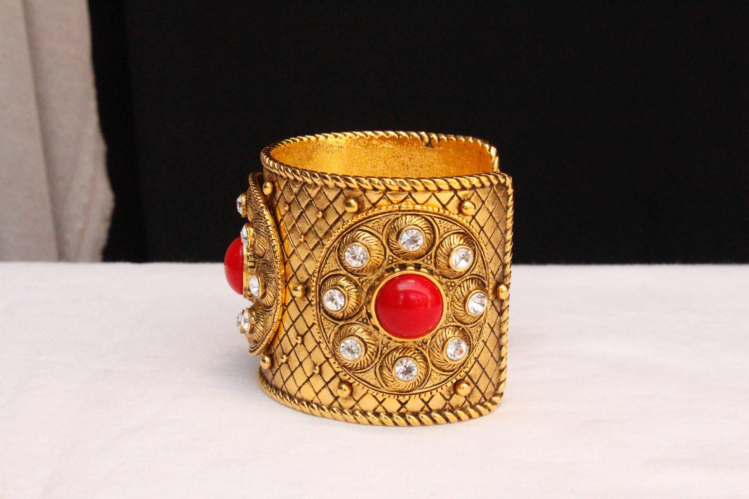CHRISTIAN DIOR BOUTIQUE – Cuff bracelet composed of chiseled gilded metal decorated with three circles paved with rhinestones and a centered red cabochon.  Signed inside.

Circa 1990.

Wrist circumference 17.5 cm (7 in); Opening 3 cm (1.25 in);