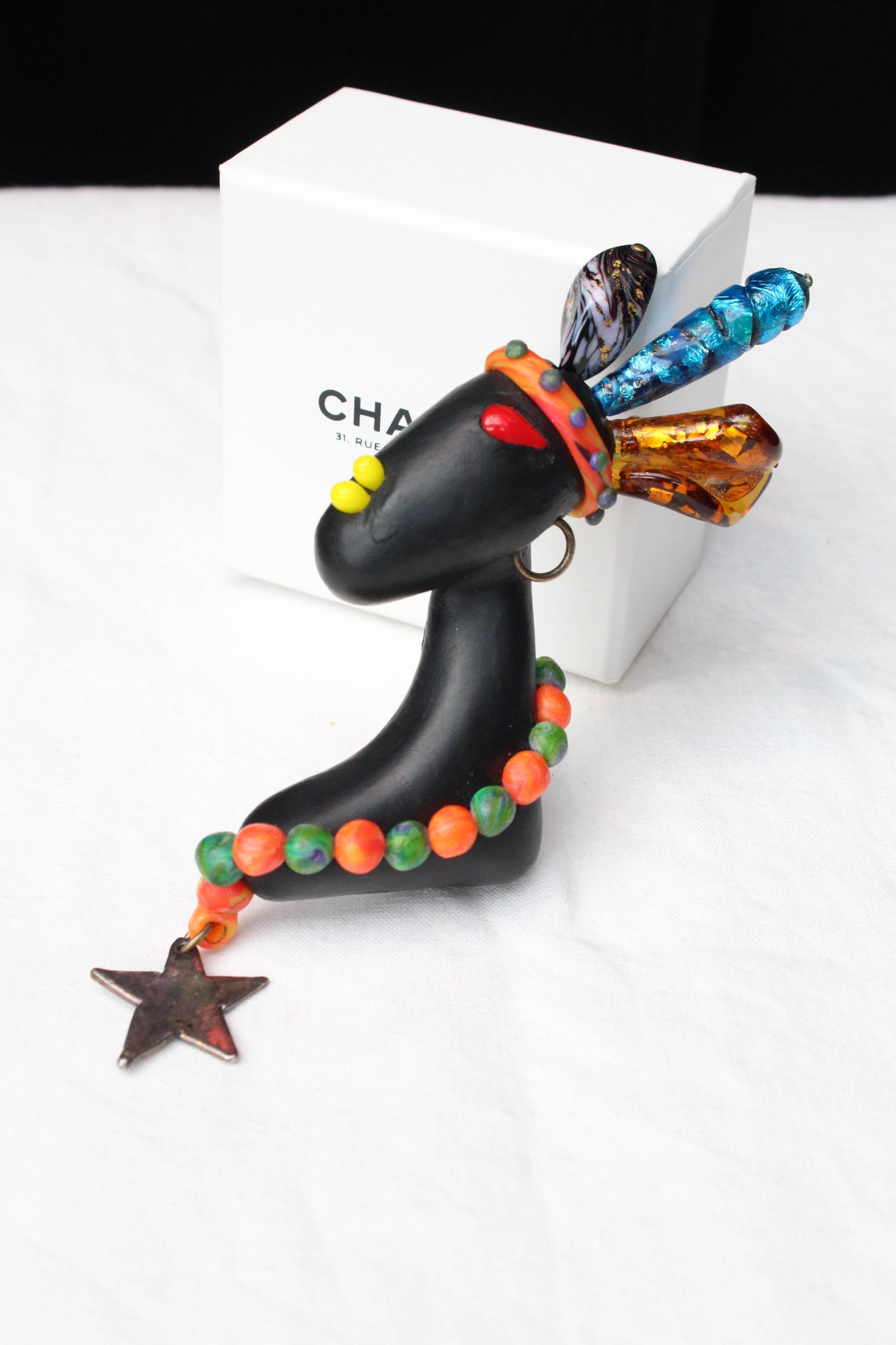 CHANEL (Made in France) Outstanding very rare polymer clay brooch representing a young African woman’s profile. The woman is wearing an orange turban topped with three Murano beads. A green and orange beads necklace finished by a silvery star