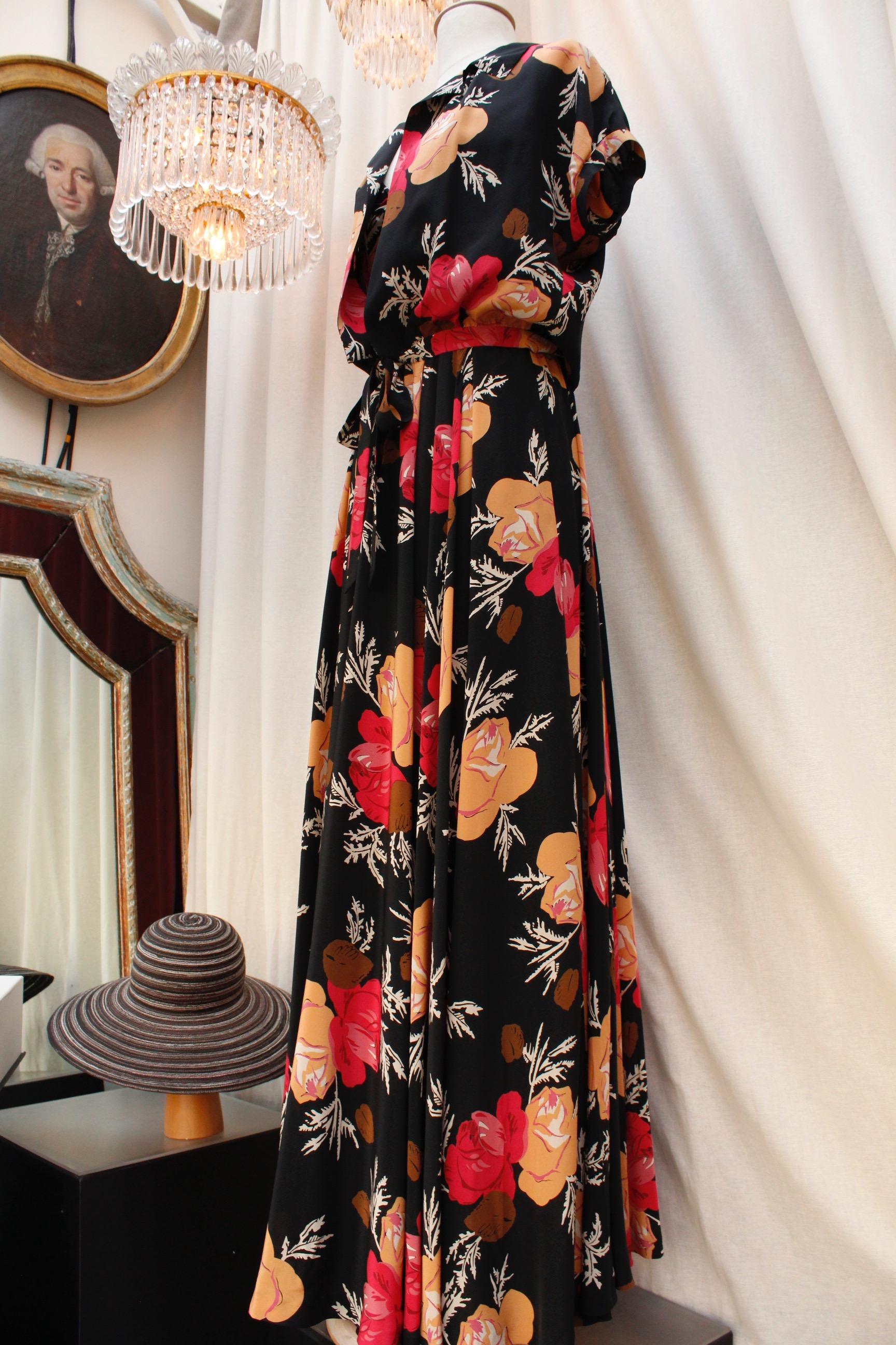 NINA RICCI (Made in France) Lovely long dress with shoulder straps, with a flared cut, a V-Neck, and pleats from the waist down. It is composed of black silk with floral pattern in orange, red, brown and white colors.  It closes with a side zip. It
