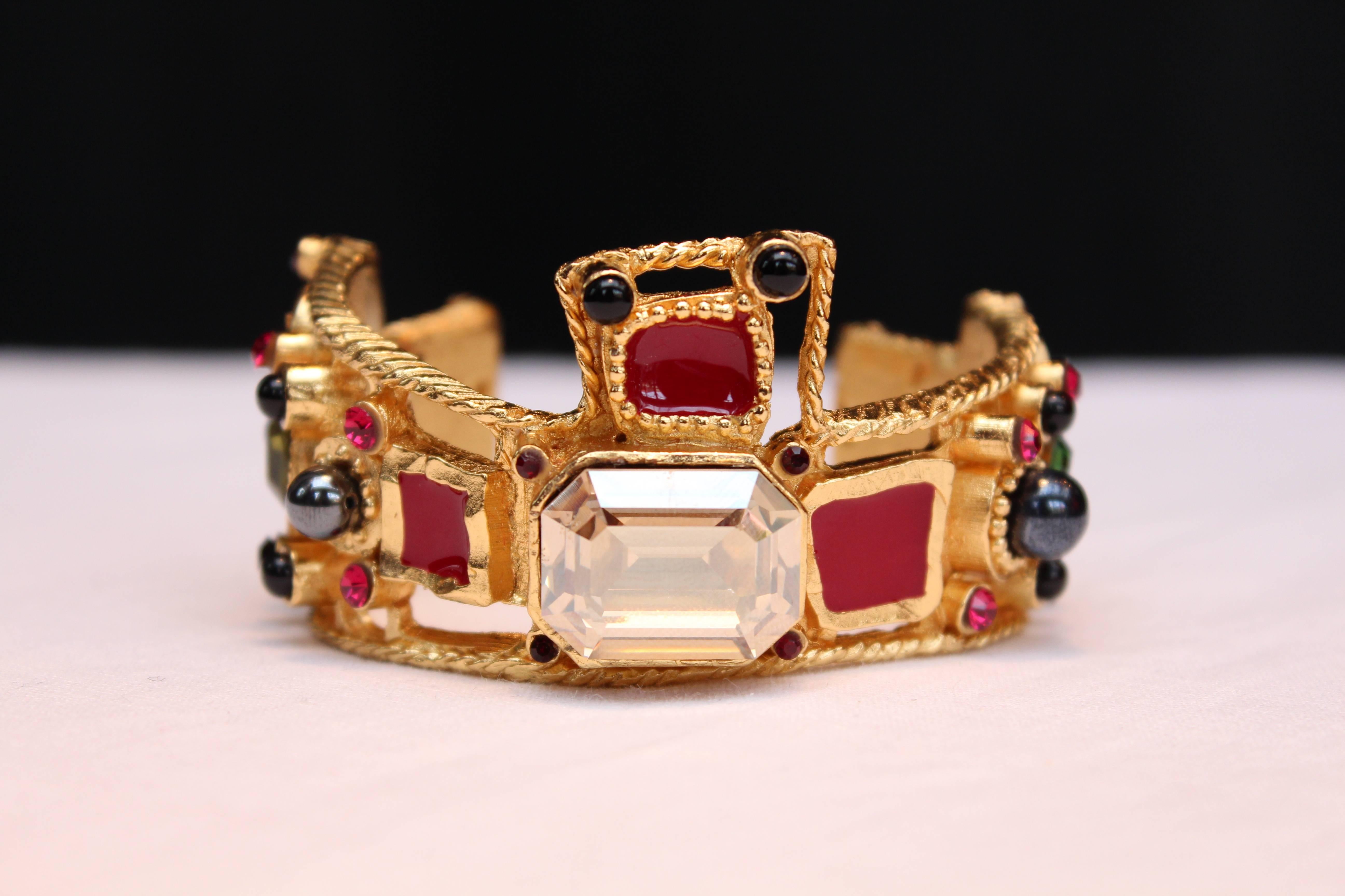 CHRISTIAN LACROIX (Made in France) Baroque cuff bracelet comprised of openwork chiseled gold metal paved with black red and blue cabochons as well green pink and white crystals. 

The cuff features in its center a large square faceted white
