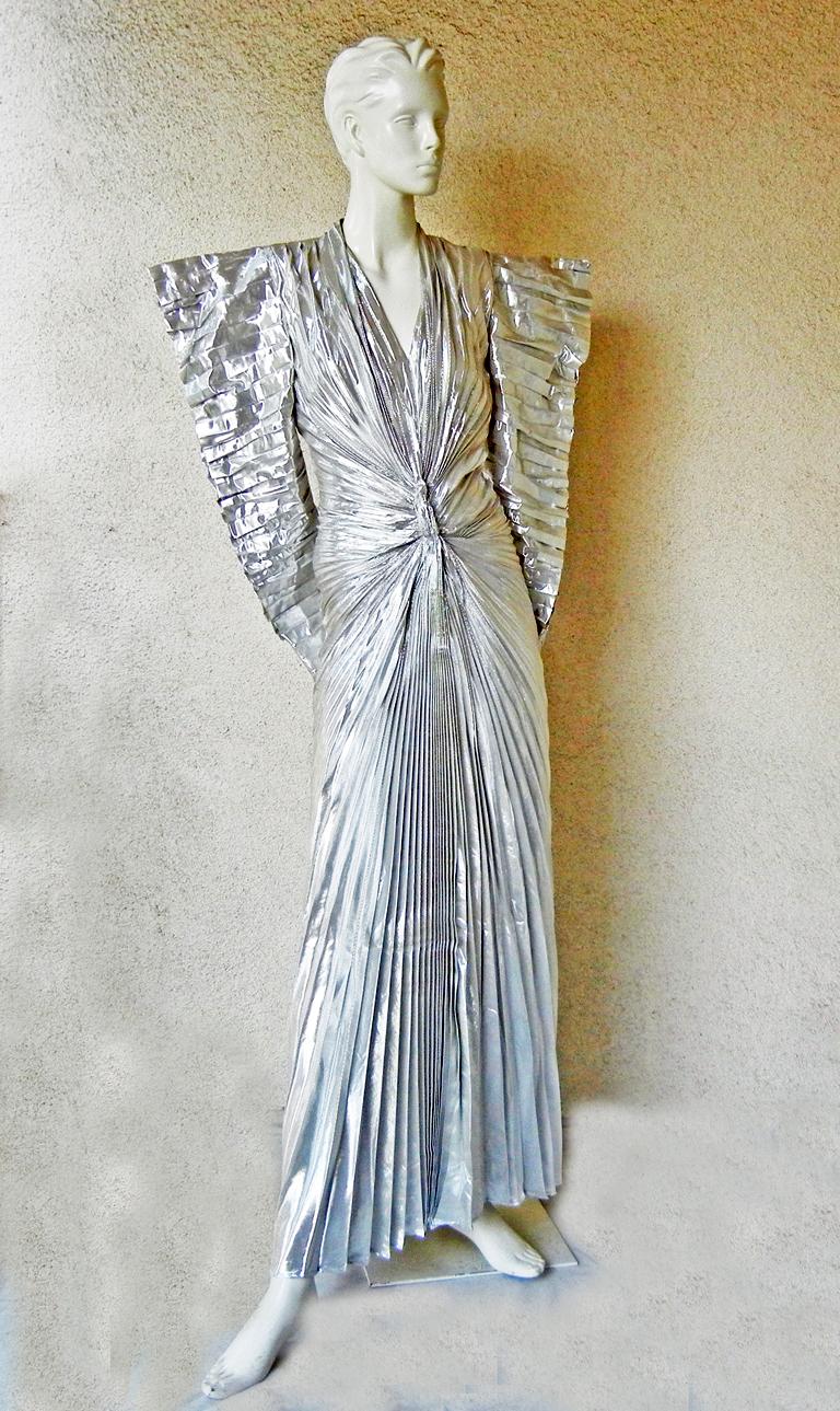 Thierry Mugler's 1979 pleated silver lame futuristic dress with wide padded stand-away shoulders, horizontally pleated sleeves with zippers, pleats converging below plunging V-neck in a sunburst design at midriff. The back is fitted over hips, then