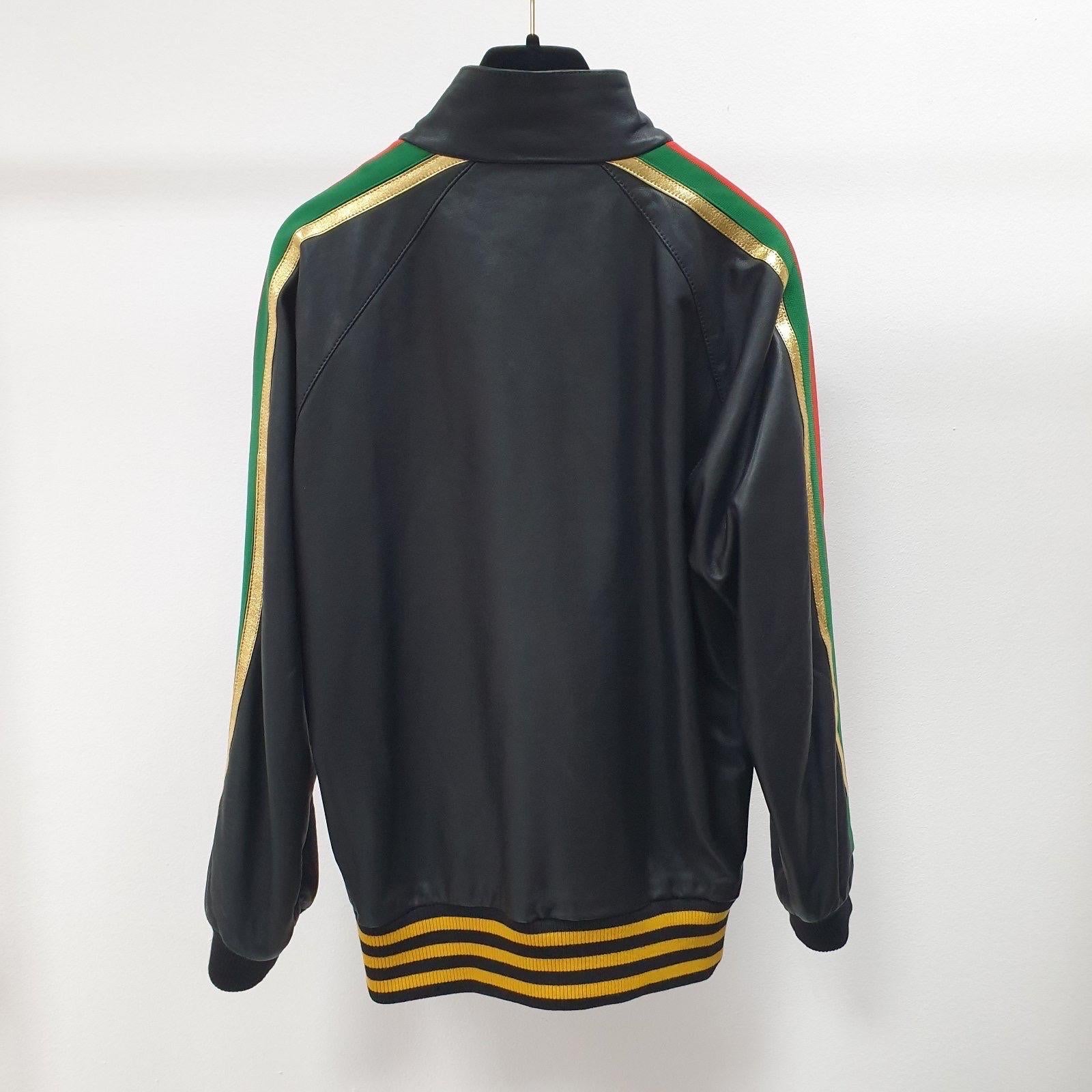 Veste de survêtement à manches longues en cuir de veau grainé noir. Col montant. Fermeture à glissière sur le devant. Logo brodé multicolore sur la poitrine. Poches passepoilées à la taille. Garniture en cuir de veau métallisé de couleur dorée et