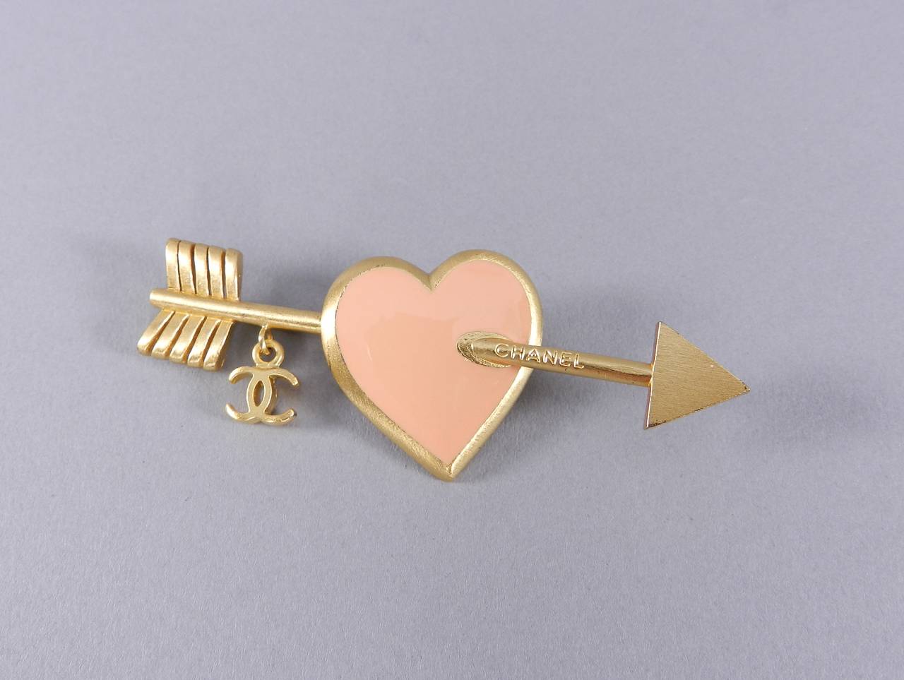 Chanel 2002 spring pink enamel heart and arrow brooch. Matte brushed goldtone metal with dangling cc charm. Excellent condition. With box. Measures 2.75 x 1 1/8