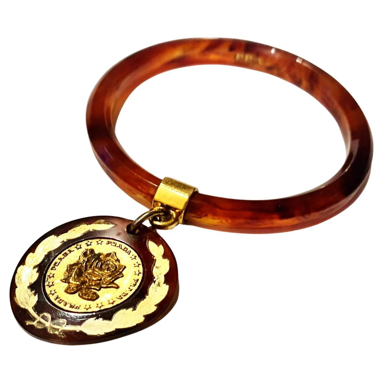 Brazalete PRADA 2000 de resina marrón, metal dorado con medallón de la marca colgando

Medidas: 8,5 cm  diámetro

Estado: años 2000, muy bueno, algunos ligeros signos de desgaste, viene con caja de marca