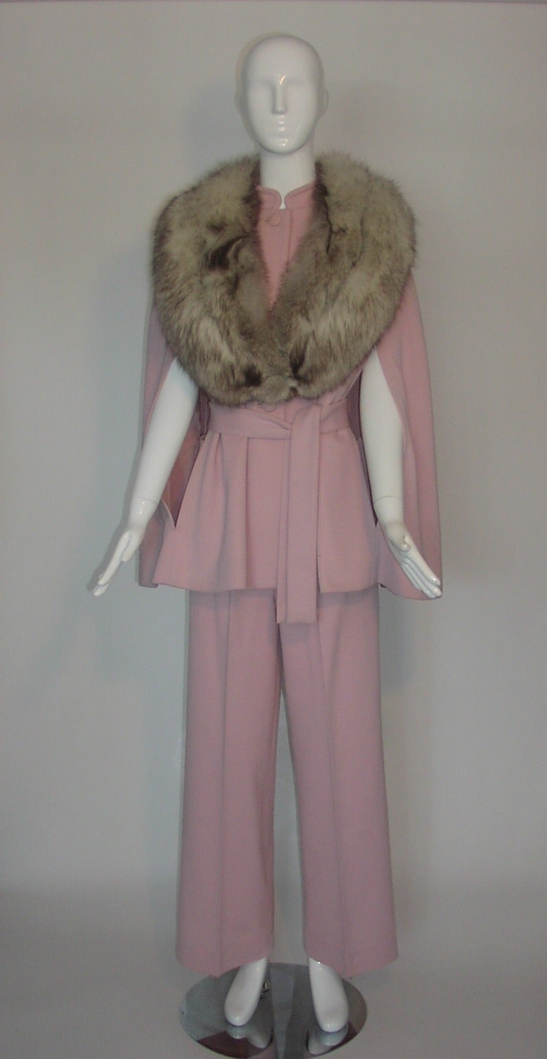 Fabulous vintage pantsuit from Lilli Ann in pink knit featuring a cape with long gray fox shawl collar. The cape has a mandarin collar, self-covered buttons, and arm slits through which a self belt may be tied; a bow with rhinestones details the