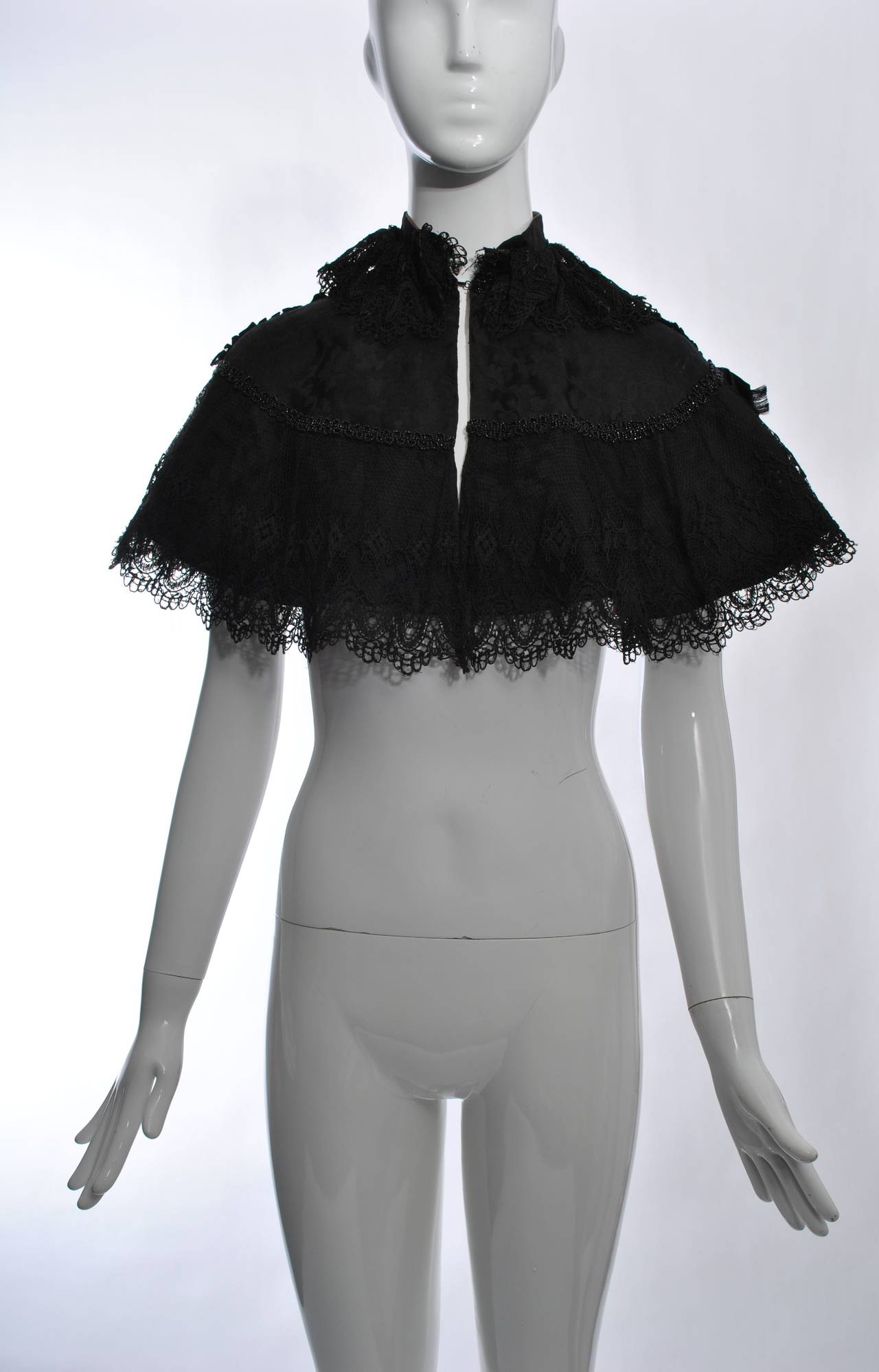 A beautiful piece to accessorize your contemporary wardrobe, this black silk capelet has a scalloped lace overlay and is embellished with a row of black beads around the upper arms and back. finished with 2 bows at the shoulders and a high-neck