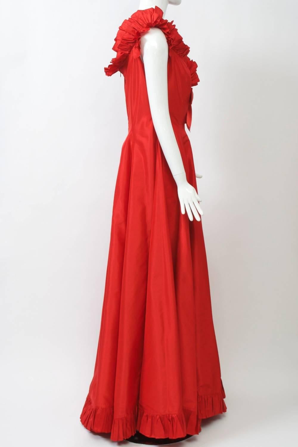 Vibrant red silk taffeta from master Oscar de la Renta shaped in princess style, sleeveless, the V-neck, hem and shoulders trimmed in a shirred ruffle and finished with a bow and rhinestone embellishment at the cleavage. Scooped and ruffled neck in