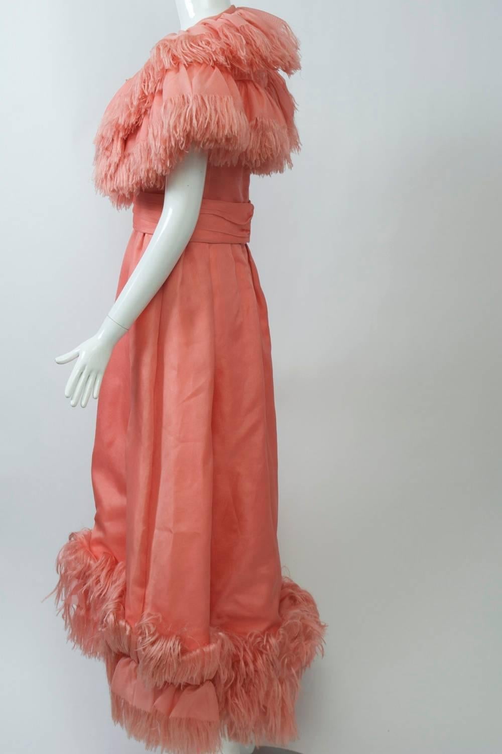 1970s custom-made  gown in peach gazar with unique fringed ruffle capelet and hem border. The sleeveless dress features a U neckline and pleated matching belt that fastens in back. Layers of the fabric comprise the double ruffle capelet, which has