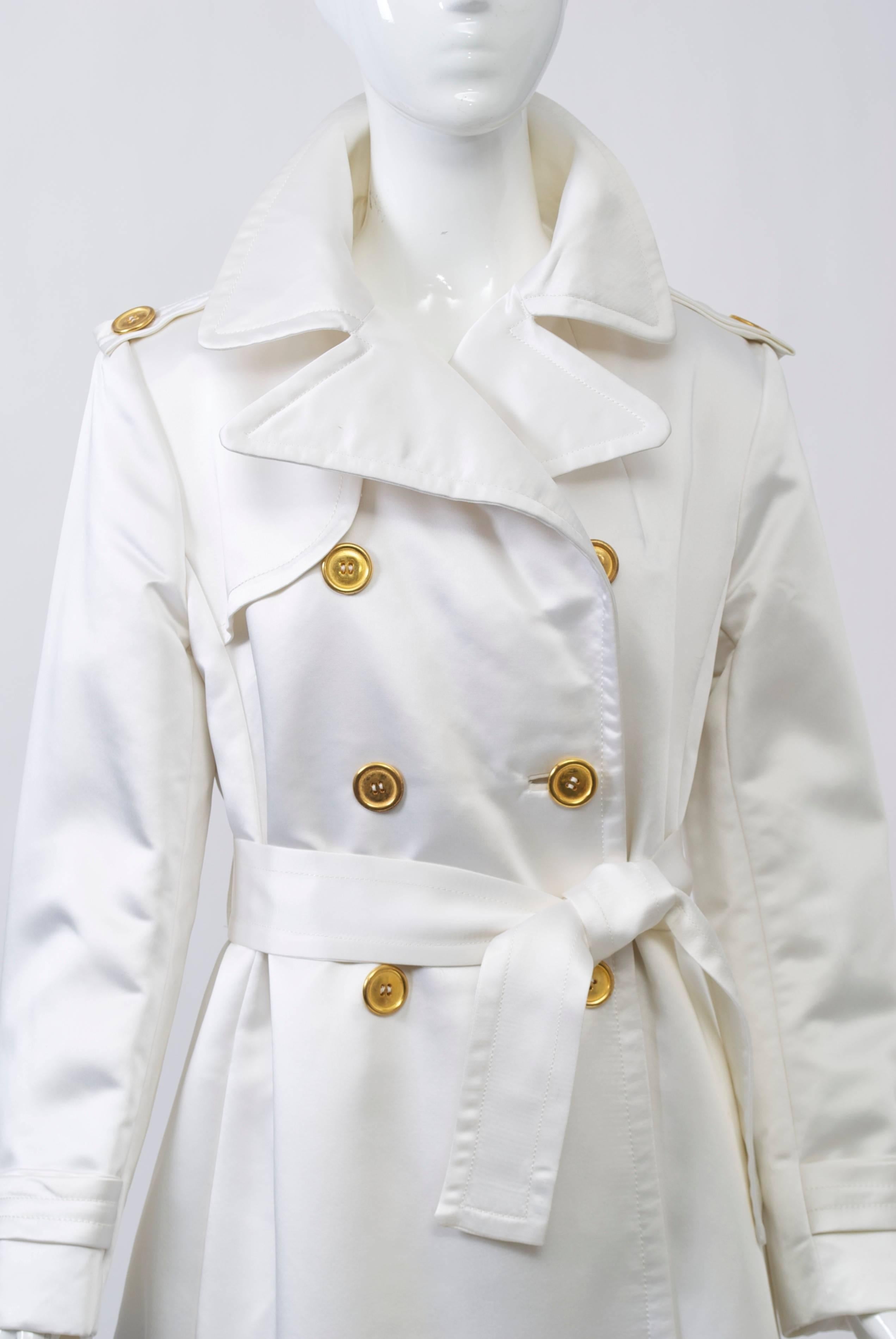 1970s long, trench-style evening coat in white satin with notched collar, epaulets, and self-tie belt. Finished with gold buttons and inverted pleat in back. Satin is backed with stiffener. 