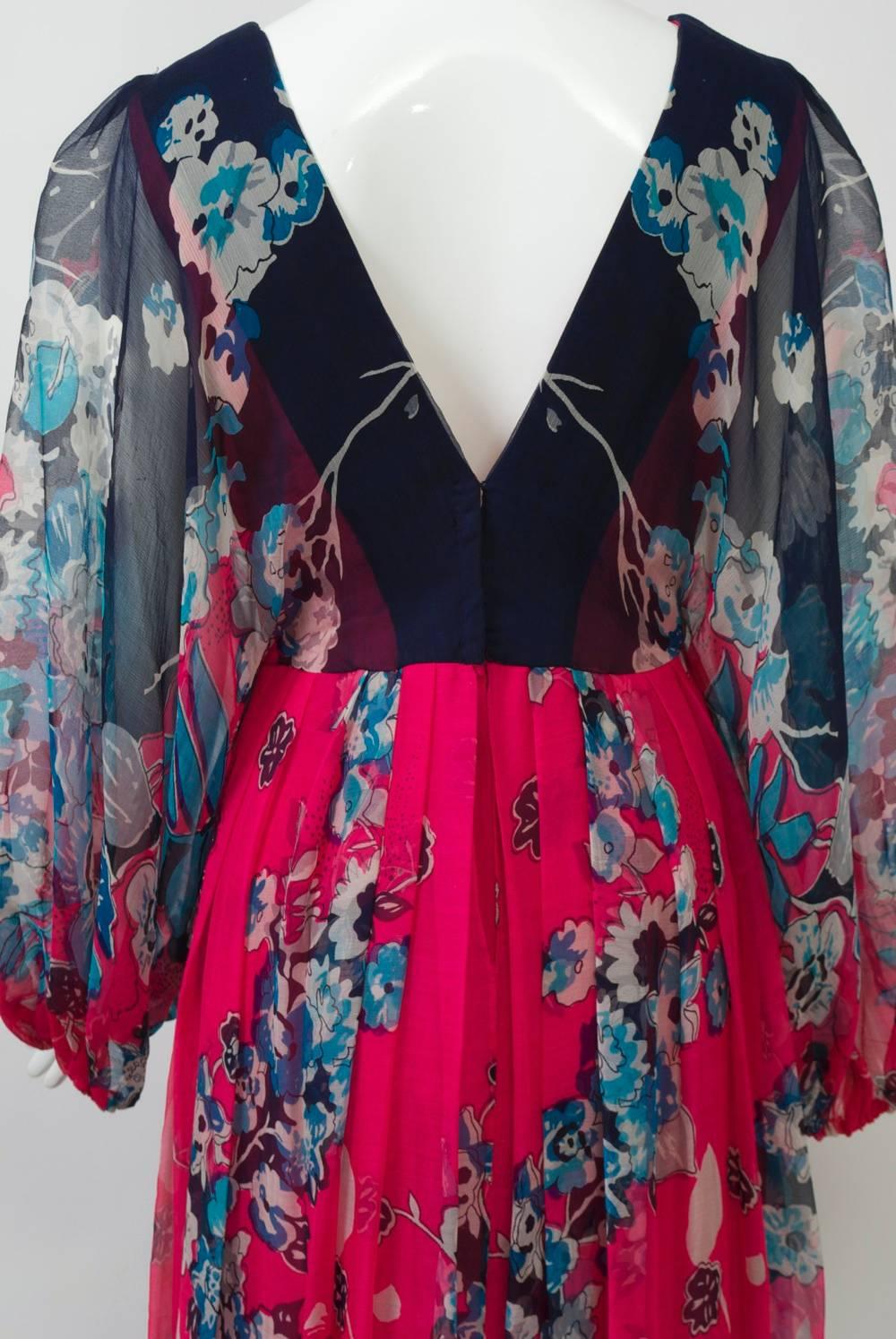 Flowing chiffon floral gown in fuchsia and navy print featuring a deep V neckline front and back, a high waistline, and sheer, raglan balloon sleeves. The skirt falls from soft pleats and has a fuchsia lining. A matching scarf can be worn around the