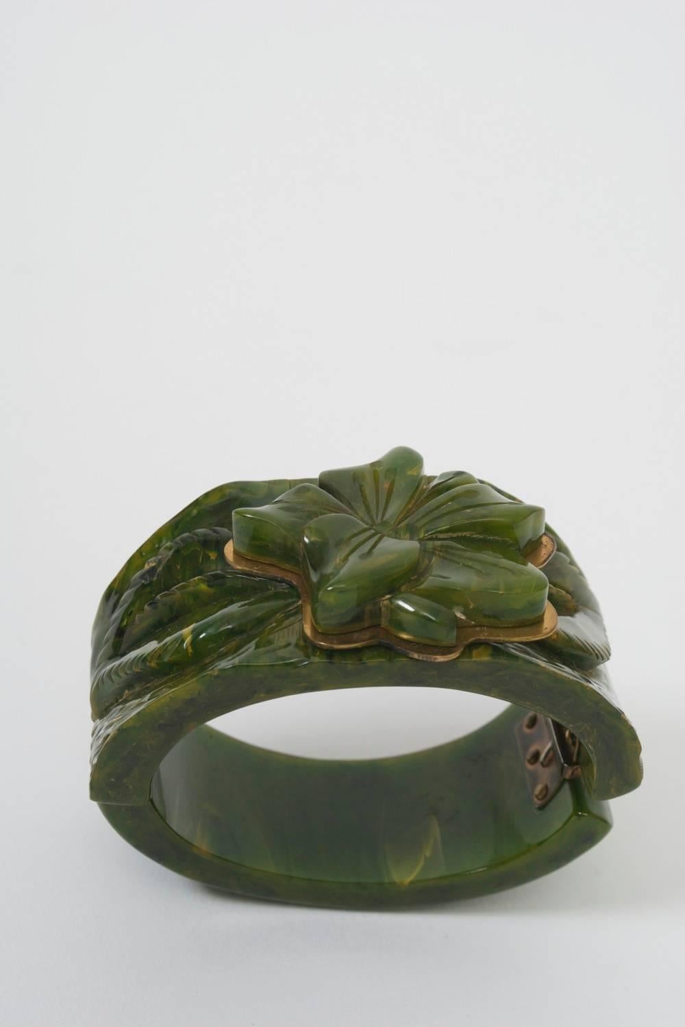 Vintage bakelite clamper bracelet in marbled green with leaf carving on the wide bangle centering an applied carved flower atop a brass cutout echoing its shape. Width at top of bangle 1.75